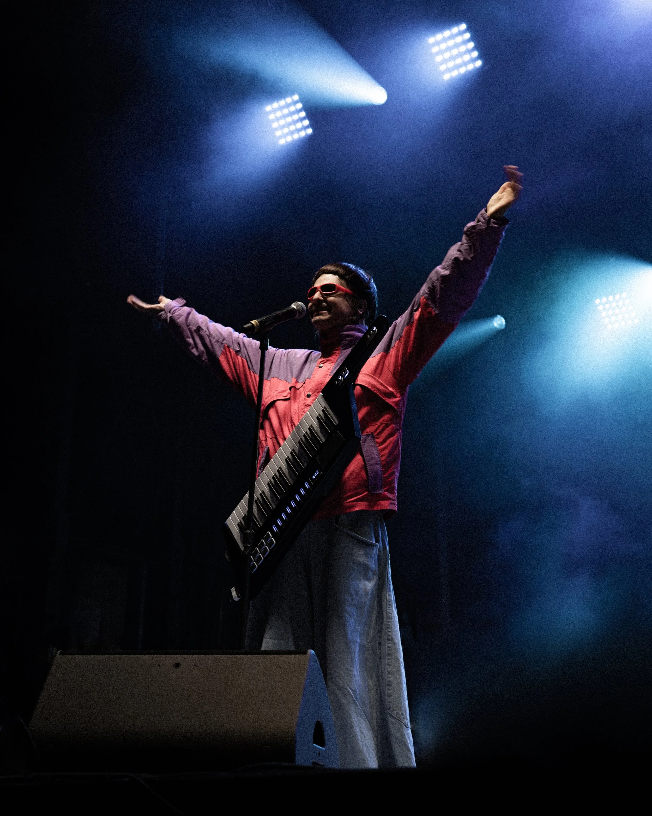 Performer on stage with arms outstretched, wearing sunglasses, a colorful jacket, and a keytar, with stage lights shining down.