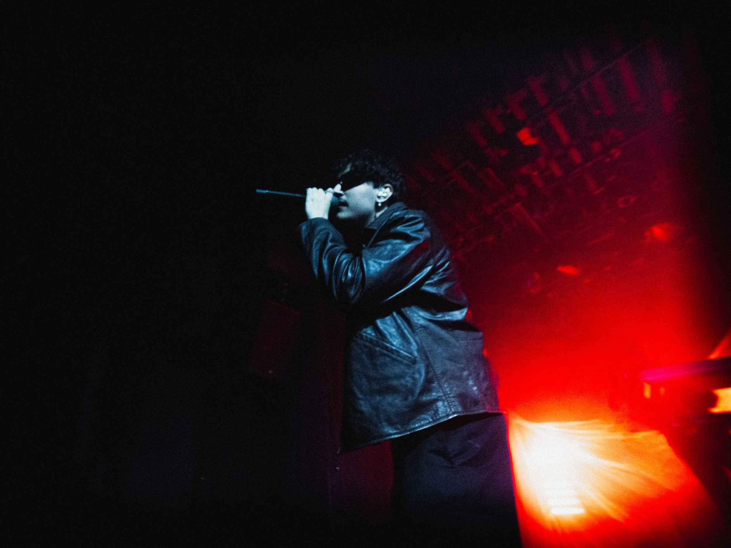 A person singing into a microphone on a stage illuminated by red and white lights, wearing a black leather jacket and dark sunglasses.