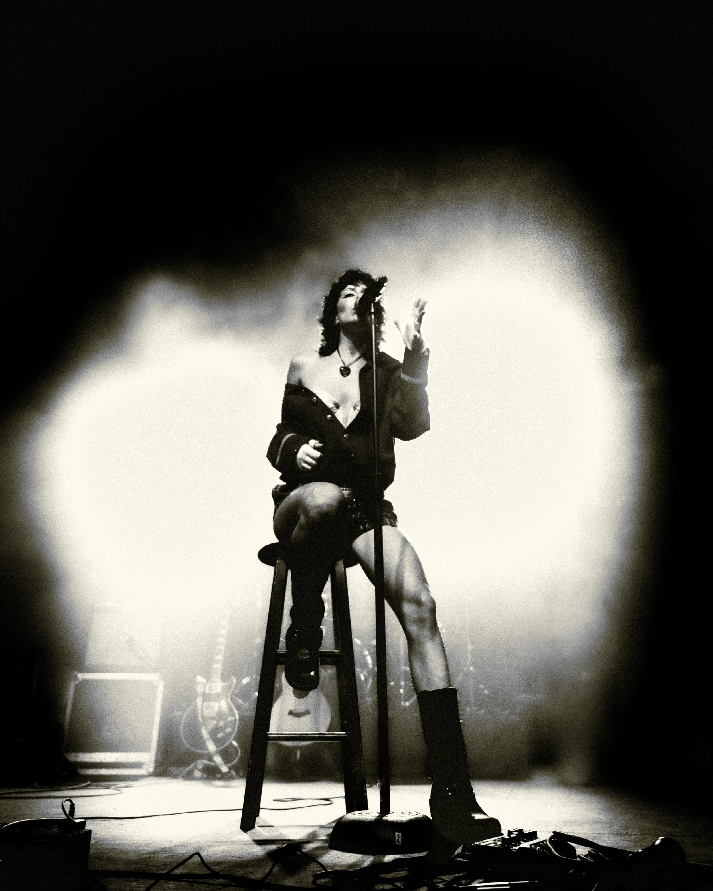 A female singer performing on stage, sitting on a stool, singing into a vintage-style microphone, with guitar and musical equipment in the background, black and white photo.