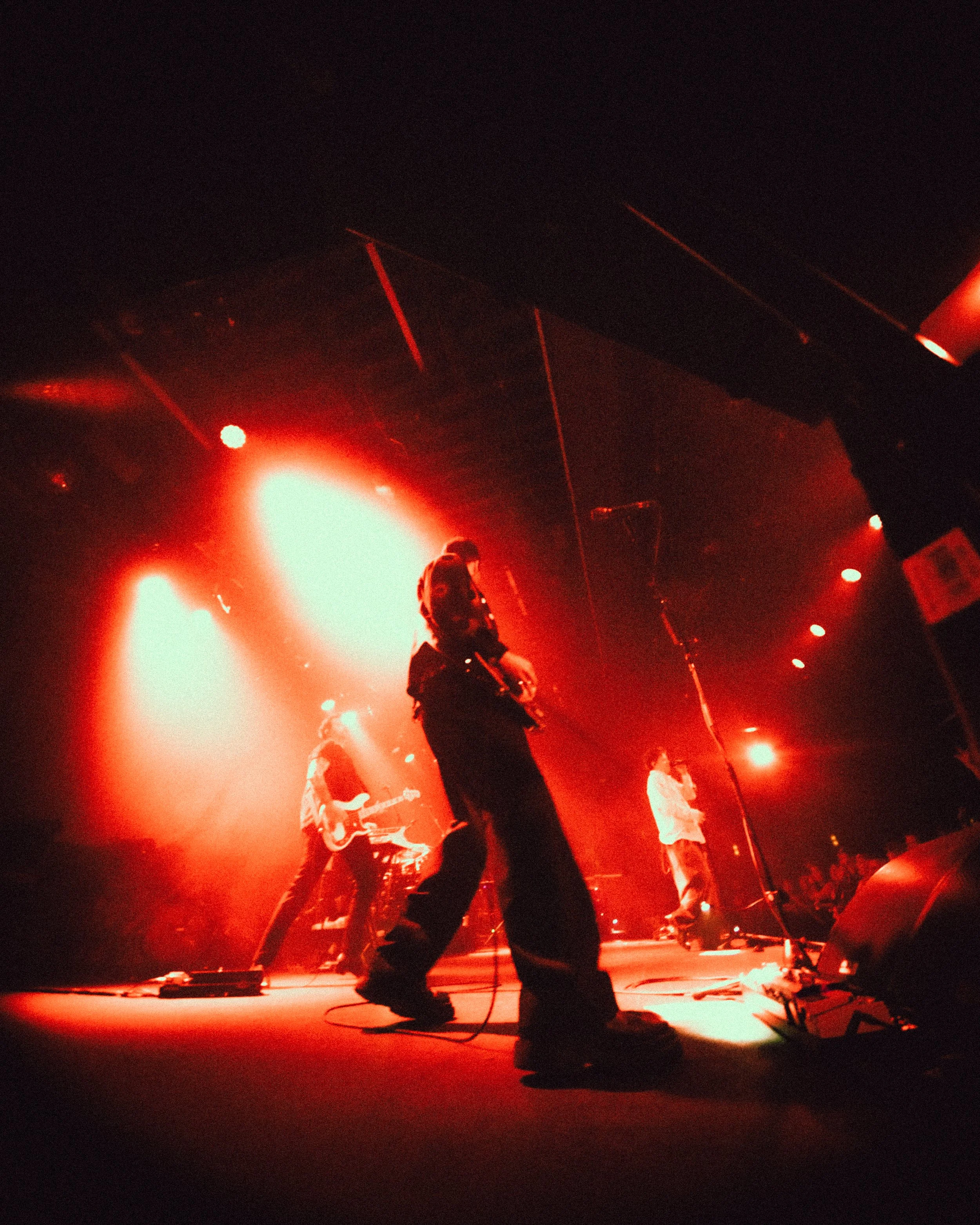 A band performing live on stage with red lighting, featuring a guitarist, a singer, and another musician, in an indoor concert venue.