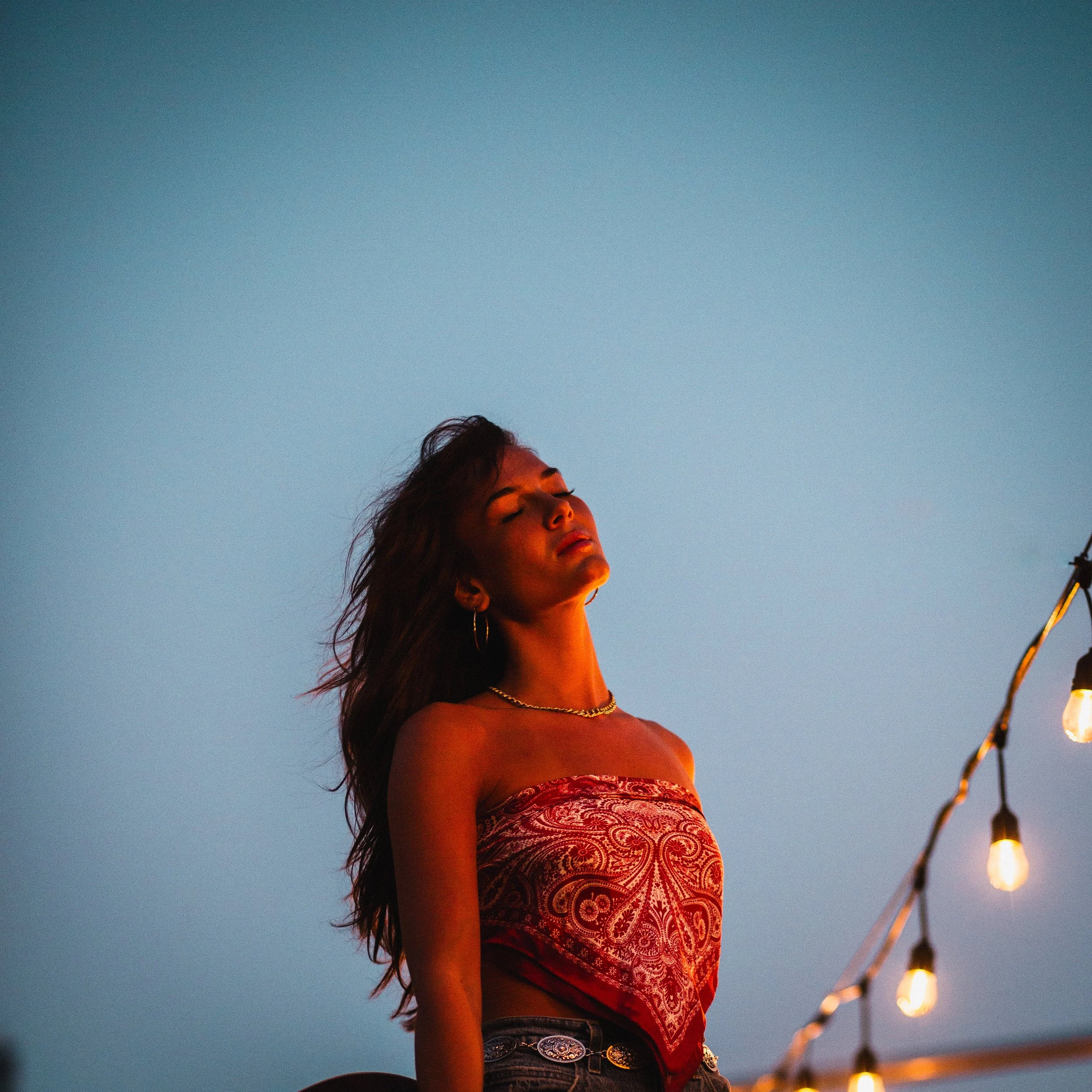 A woman with long dark hair, eyes closed, wearing a red patterned tube top and jewelry, standing outdoors during sunset with string lights nearby.