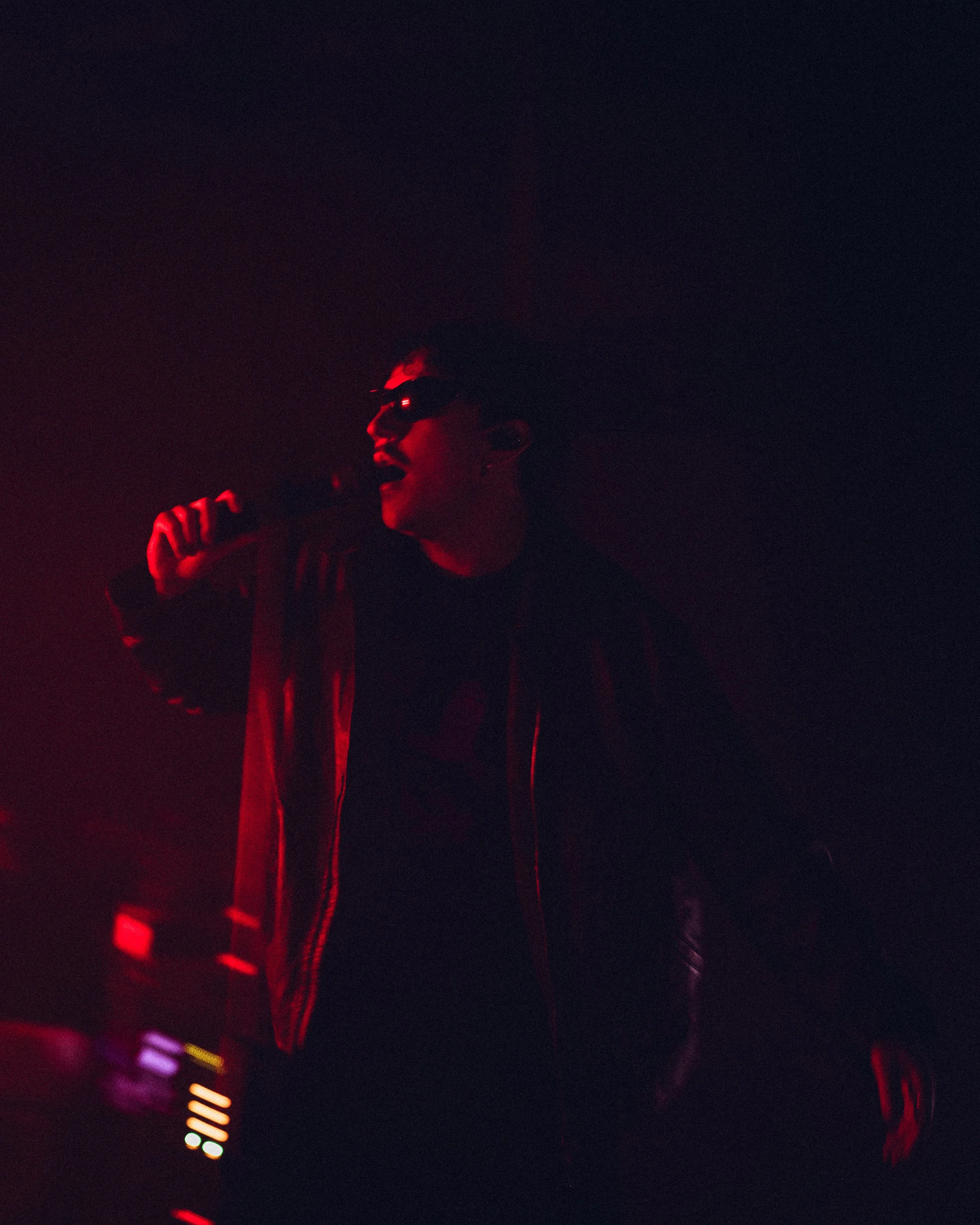 A person wearing sunglasses and a leather jacket holding a microphone in a dark environment with red lighting.