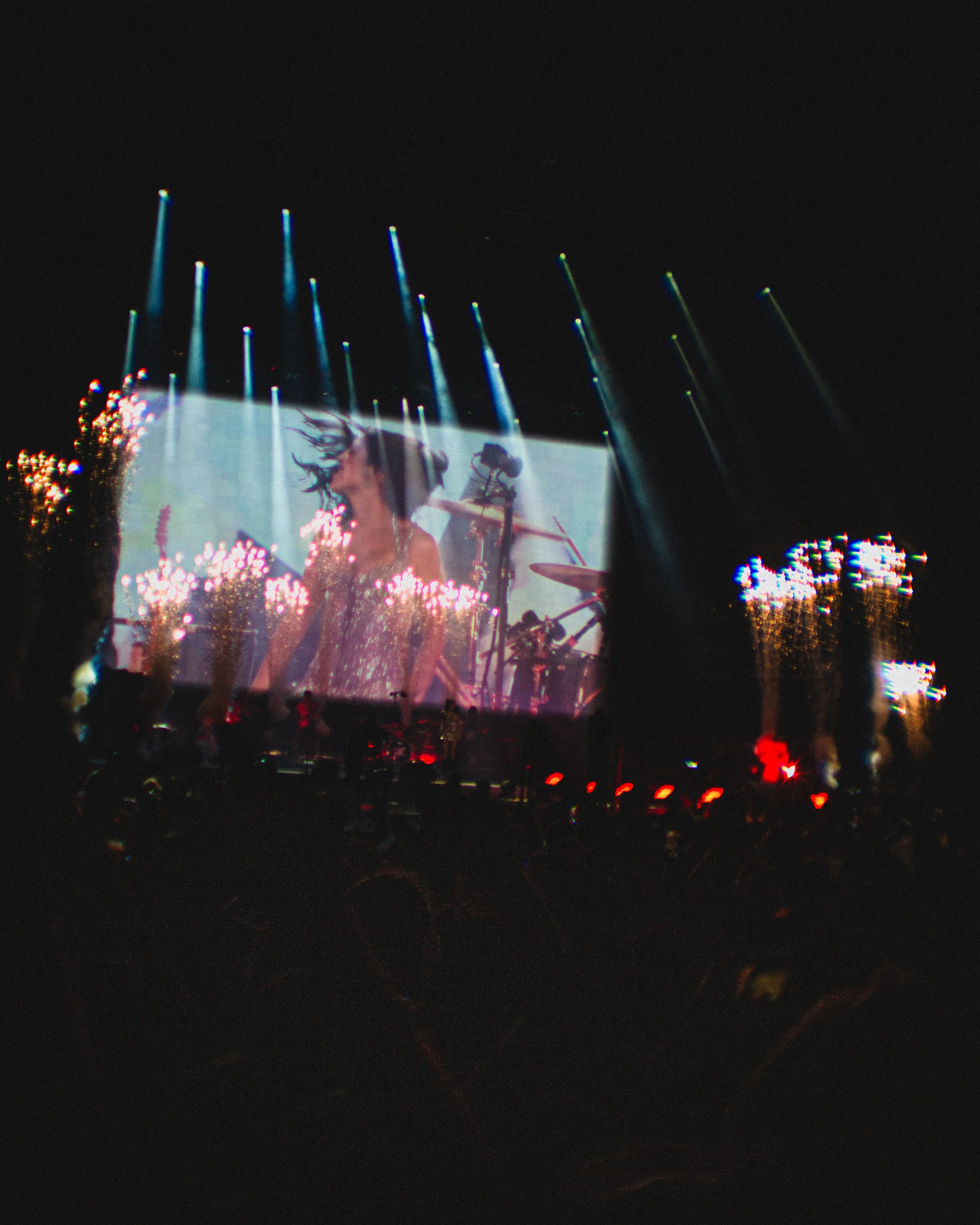 A concert scene showing a large screen with a performer, stage lights, and smoke effects at night.