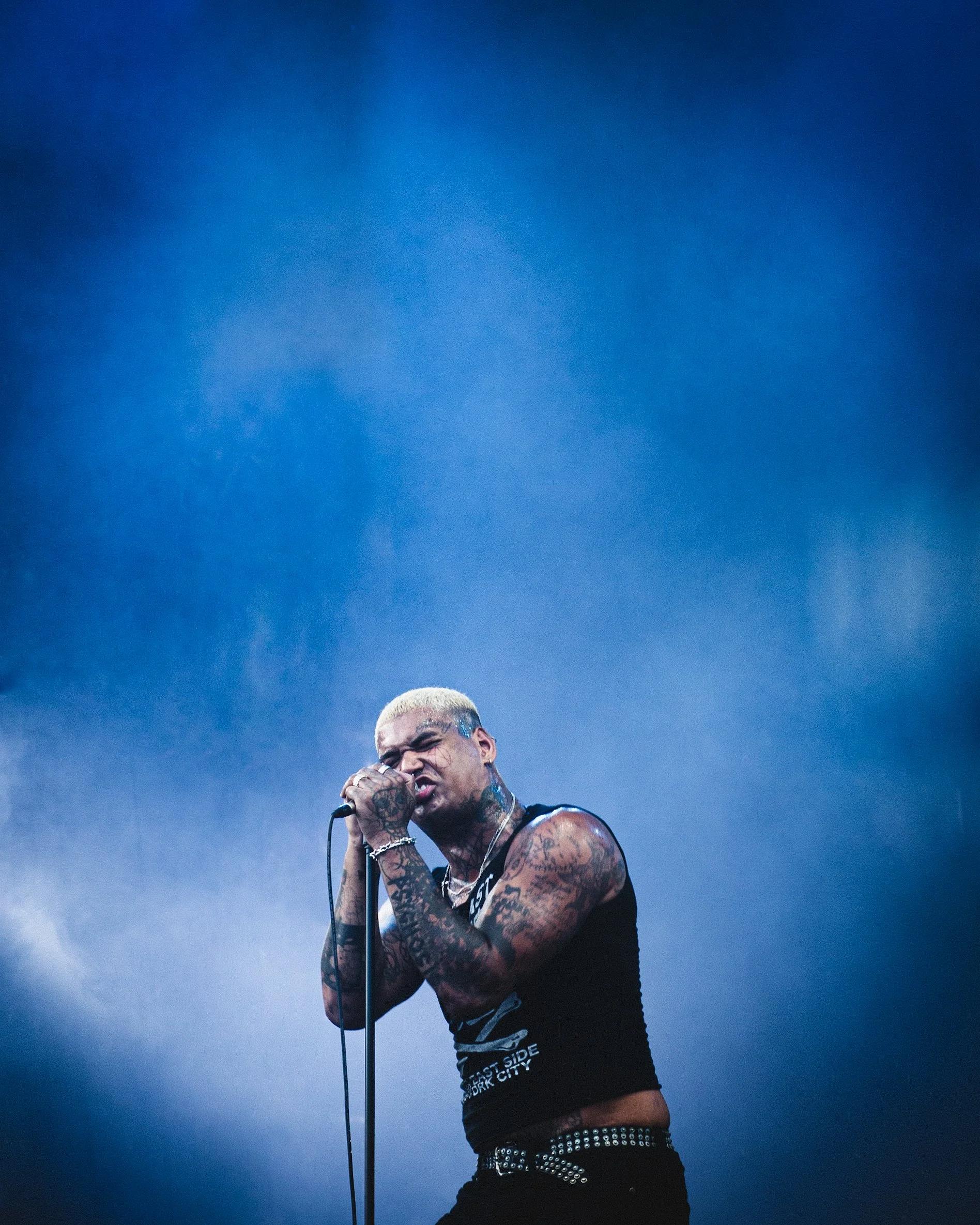A tattooed man with blond hair passionately singing into a microphone on stage against a blue background.