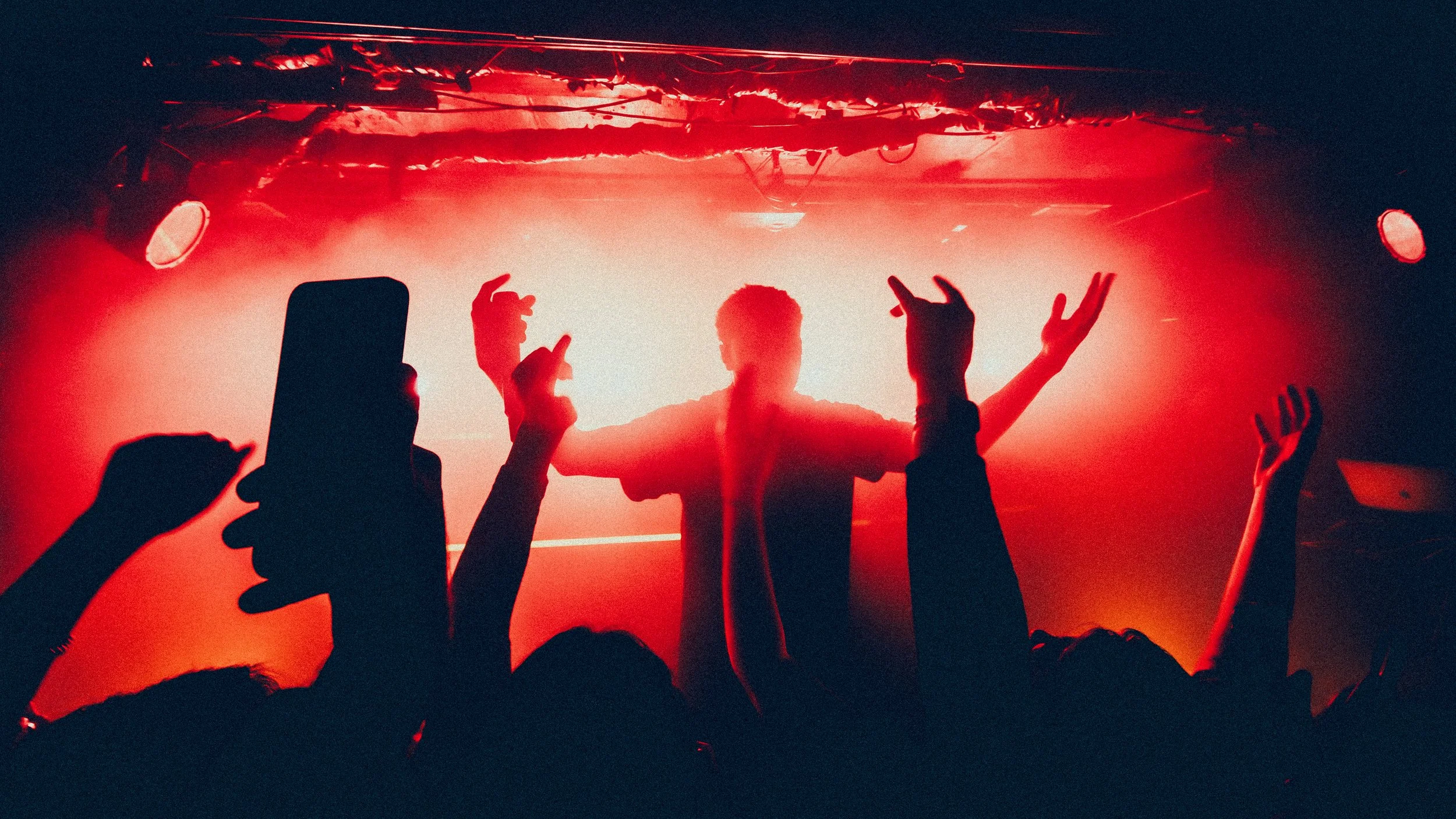 Silhouette of a person dancing on stage with arms raised, backlit by a bright red and orange light, with an audience in front holding up phones and hands.