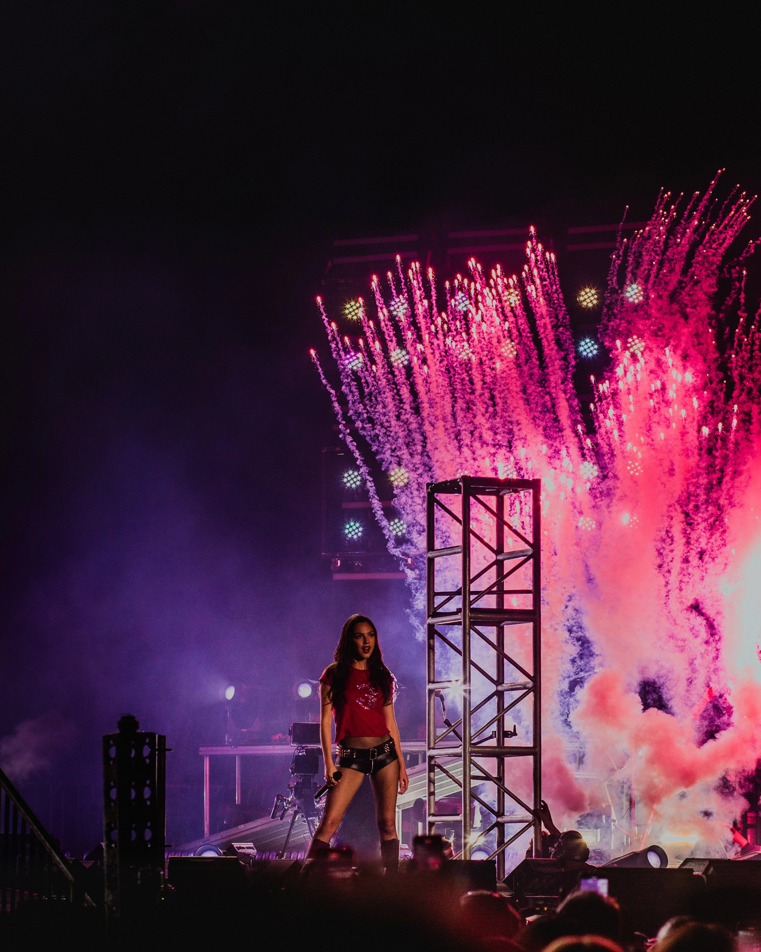 A woman with long dark hair in a red crop top and black shorts standing on a stage during a fireworks display at night.