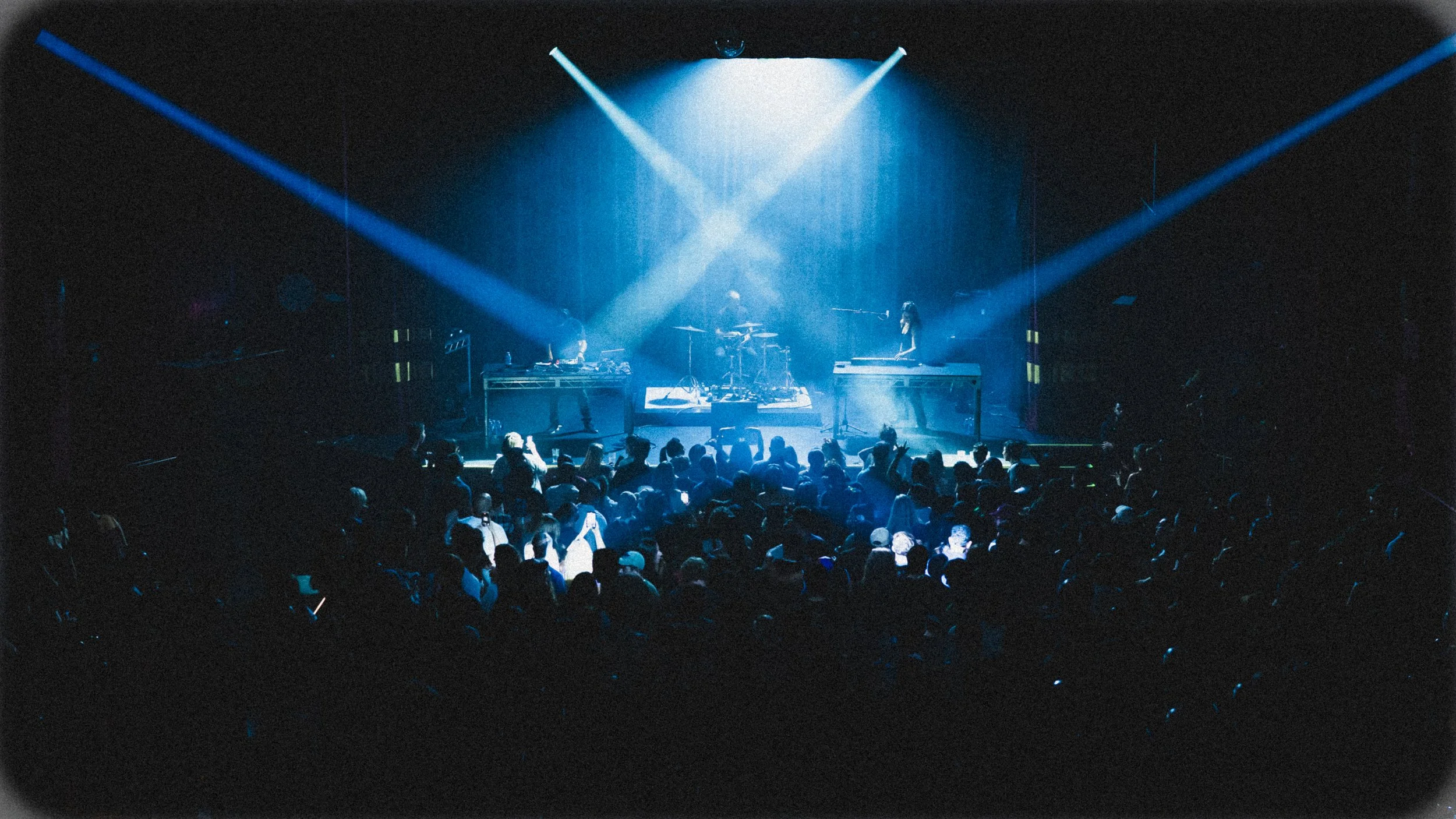 A concert stage with blue lighting and two musicians playing keyboards, with a crowd of people dancing and enjoying the music.