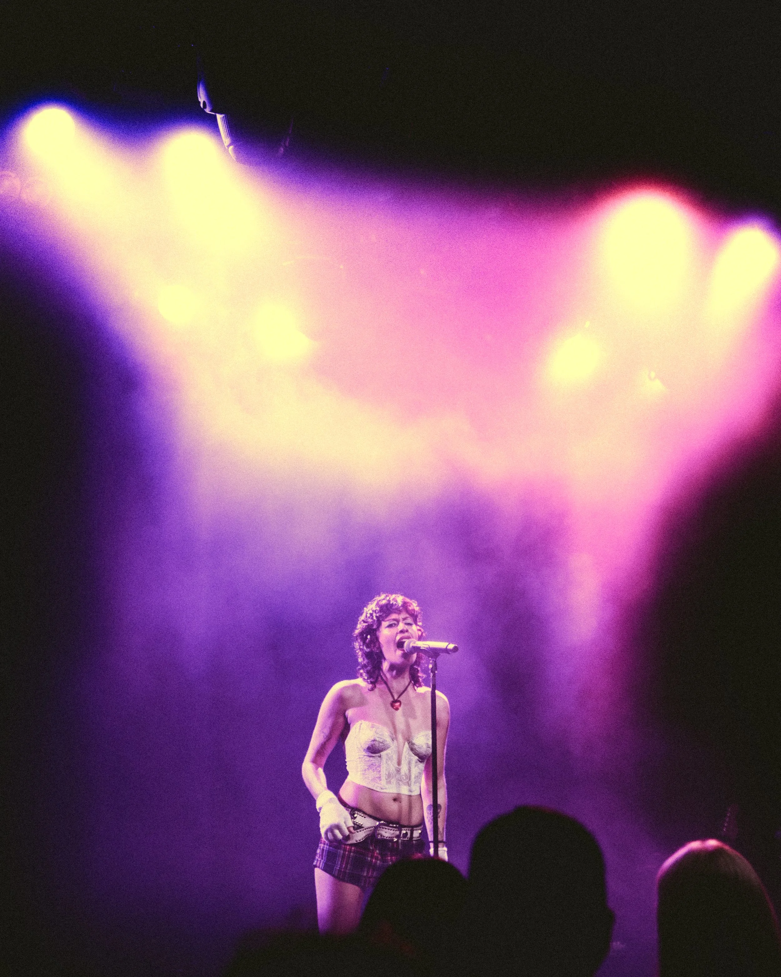 A woman singing on stage with colorful purple and pink lighting, wearing a sleeveless top and shorts, with an audience in the foreground.