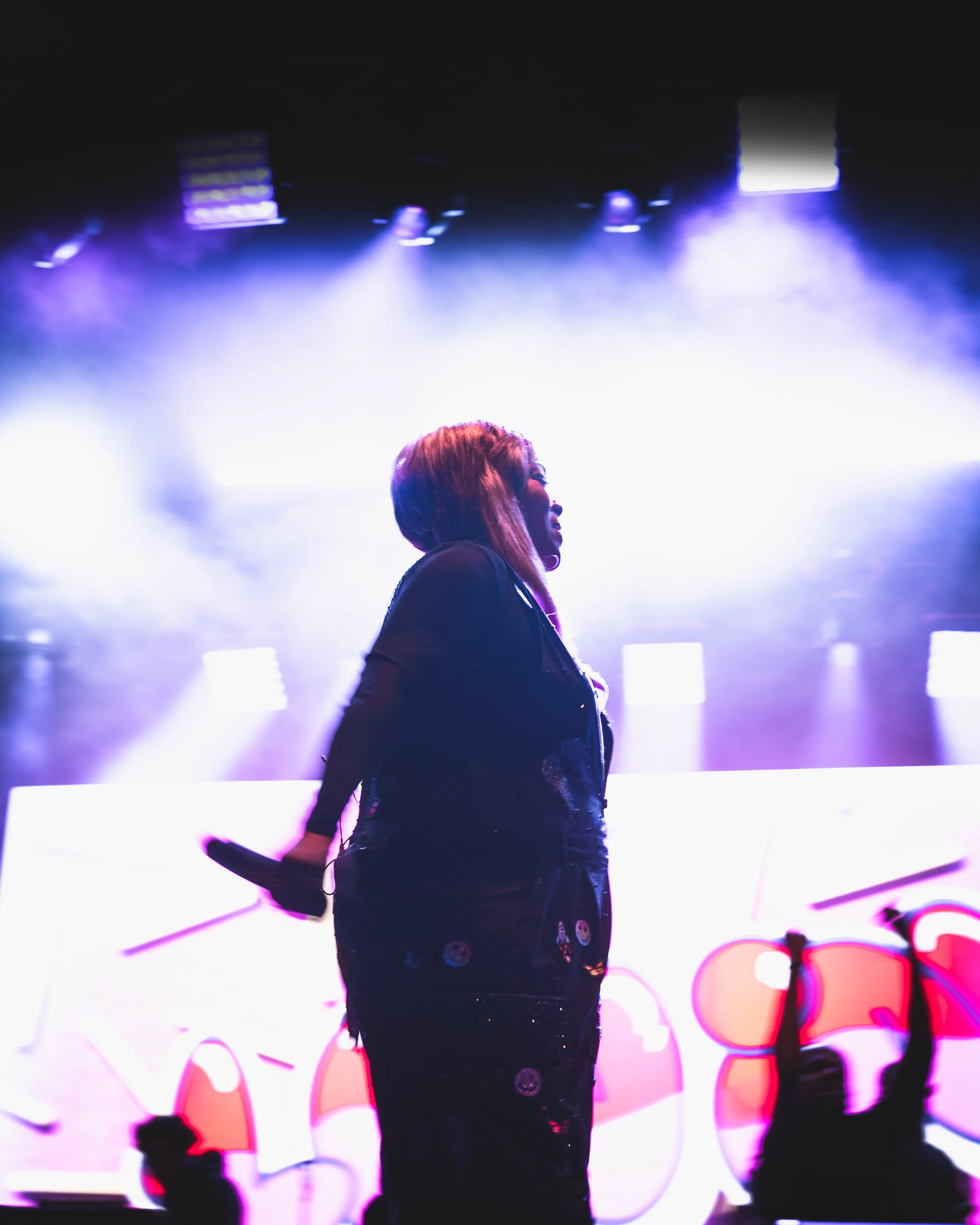 A female performer on stage with purple and white lighting, holding a microphone behind her back, and raising her fist in the air.