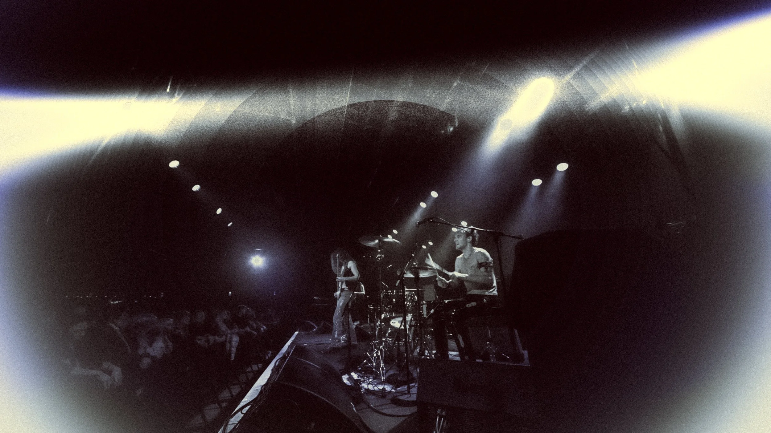 Musicians performing on stage with drums and other instruments, audience sitting in front, dark ambiance with spotlights.
