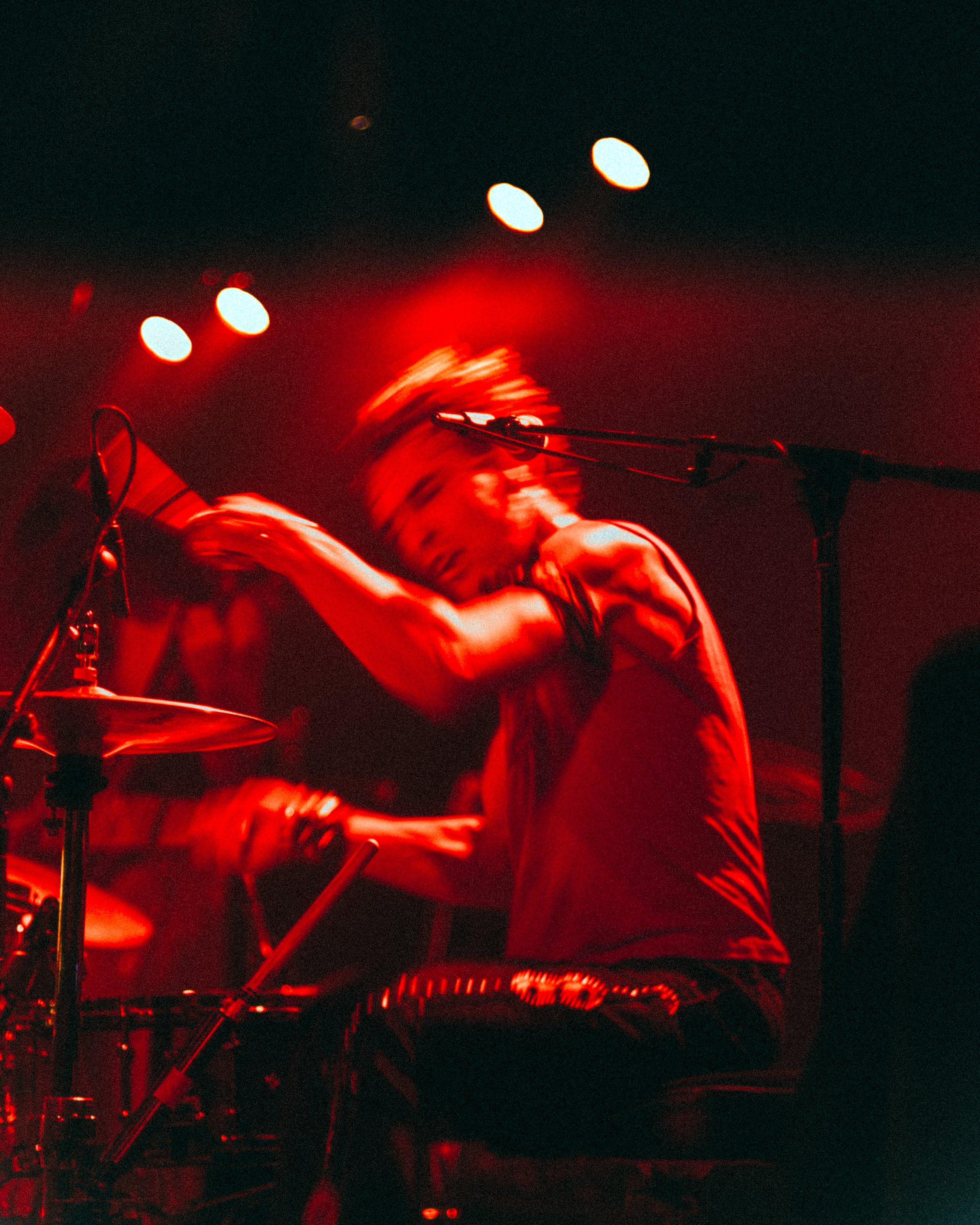 A person performing on stage in red lighting, playing drums with motion blur indicating energetic movement.