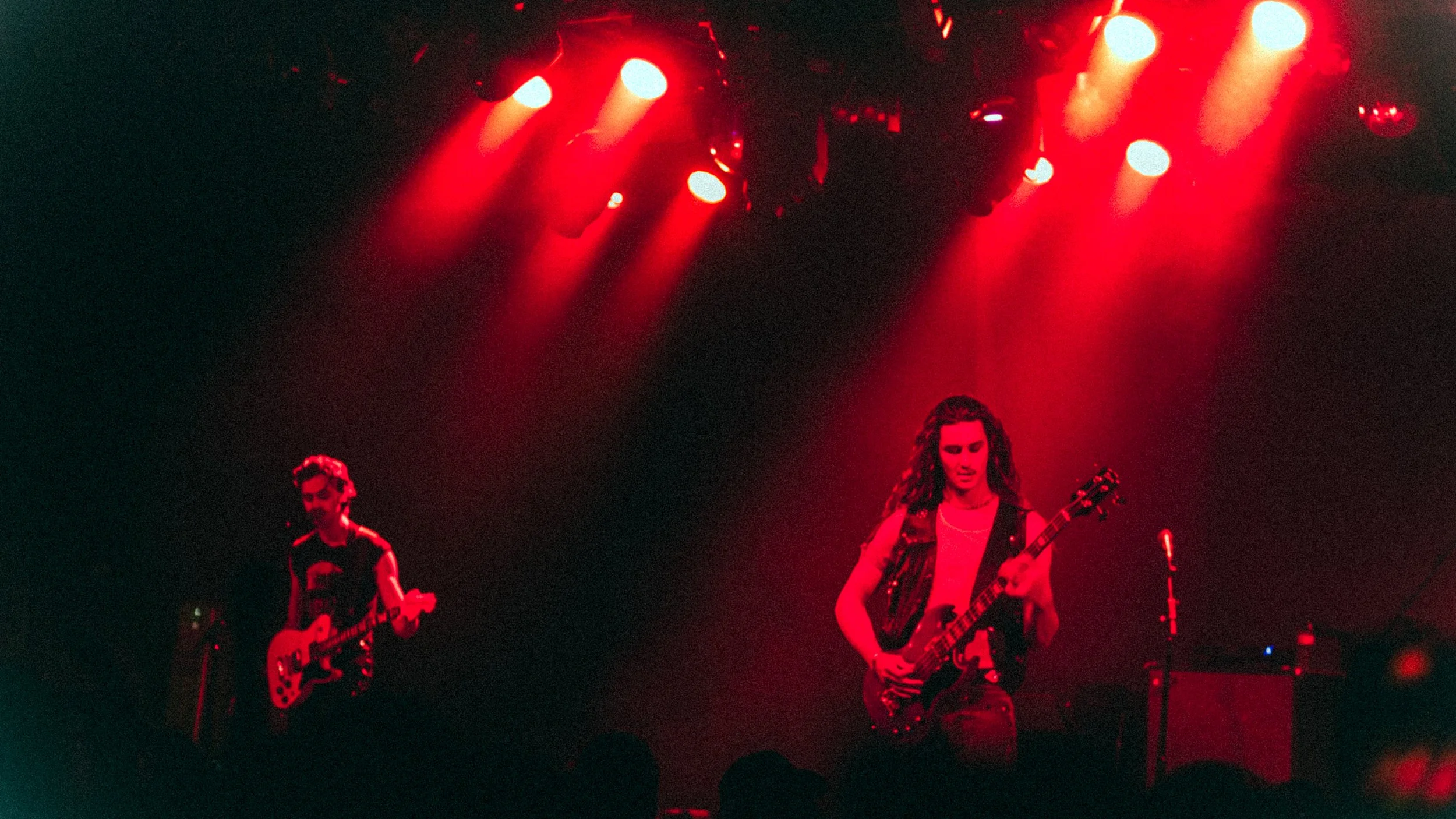 Musicians performing on stage under red lighting; one person playing guitar on the right, another playing guitar on the left.