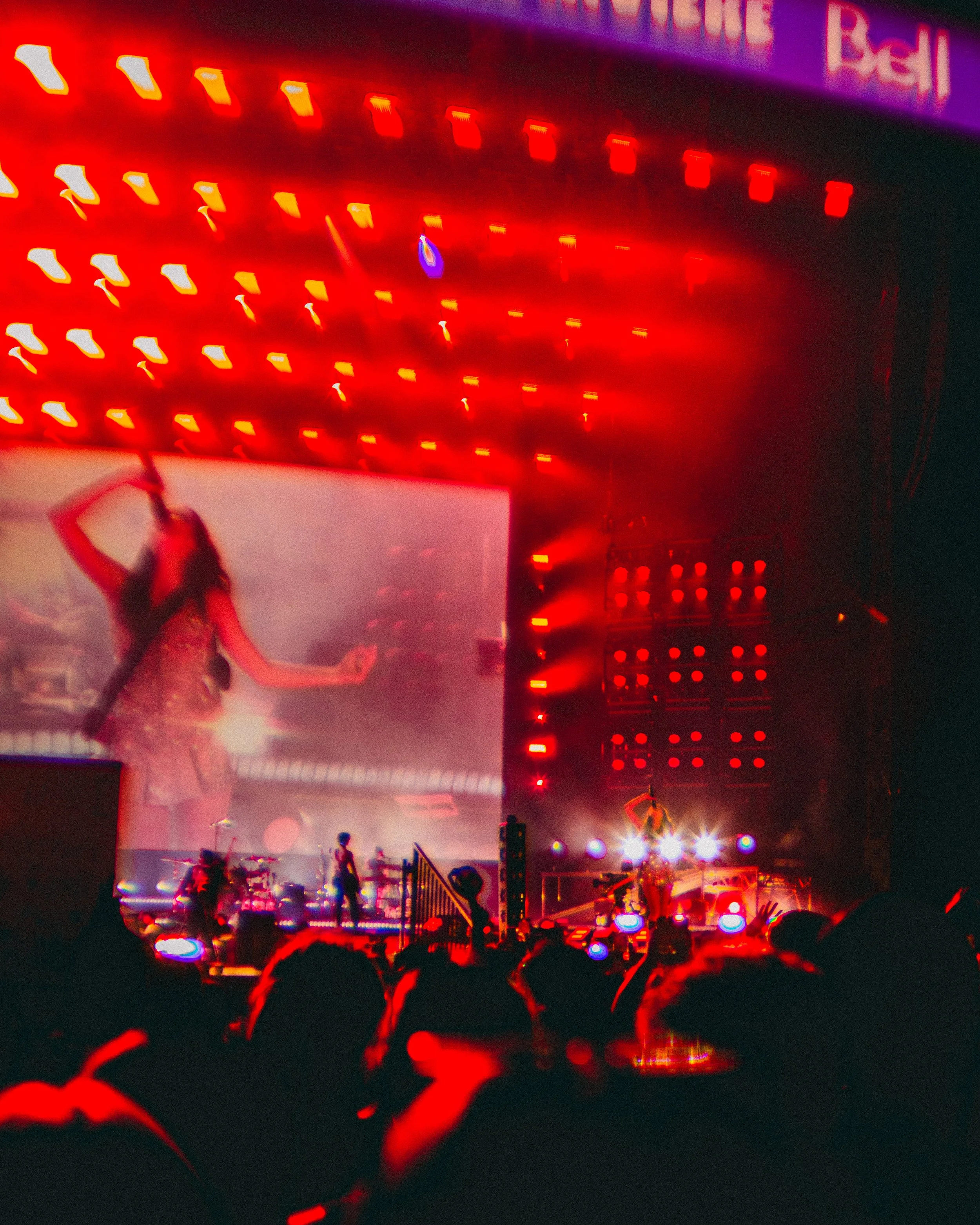 Concert stage with bright red and white lights, performers with musical instruments, and a large screen projecting an image of a woman with long hair in a dress. Audience members are visible at the bottom.