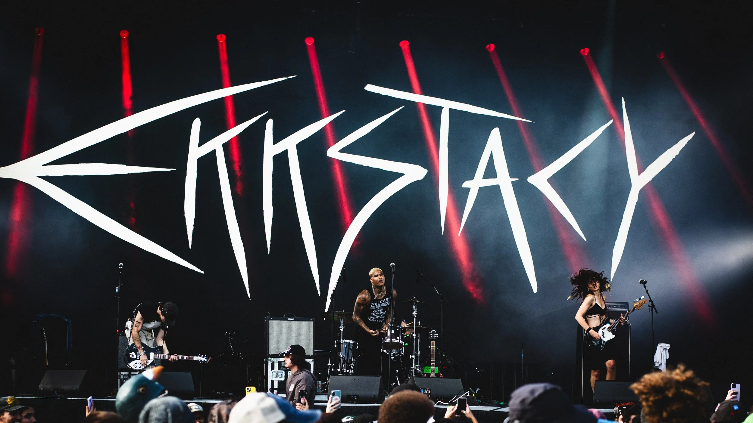 A live concert with a band performing on stage, with a large illuminated sign that reads "KICKSKY" in the background, red spotlights shining above, and audience members taking photos.