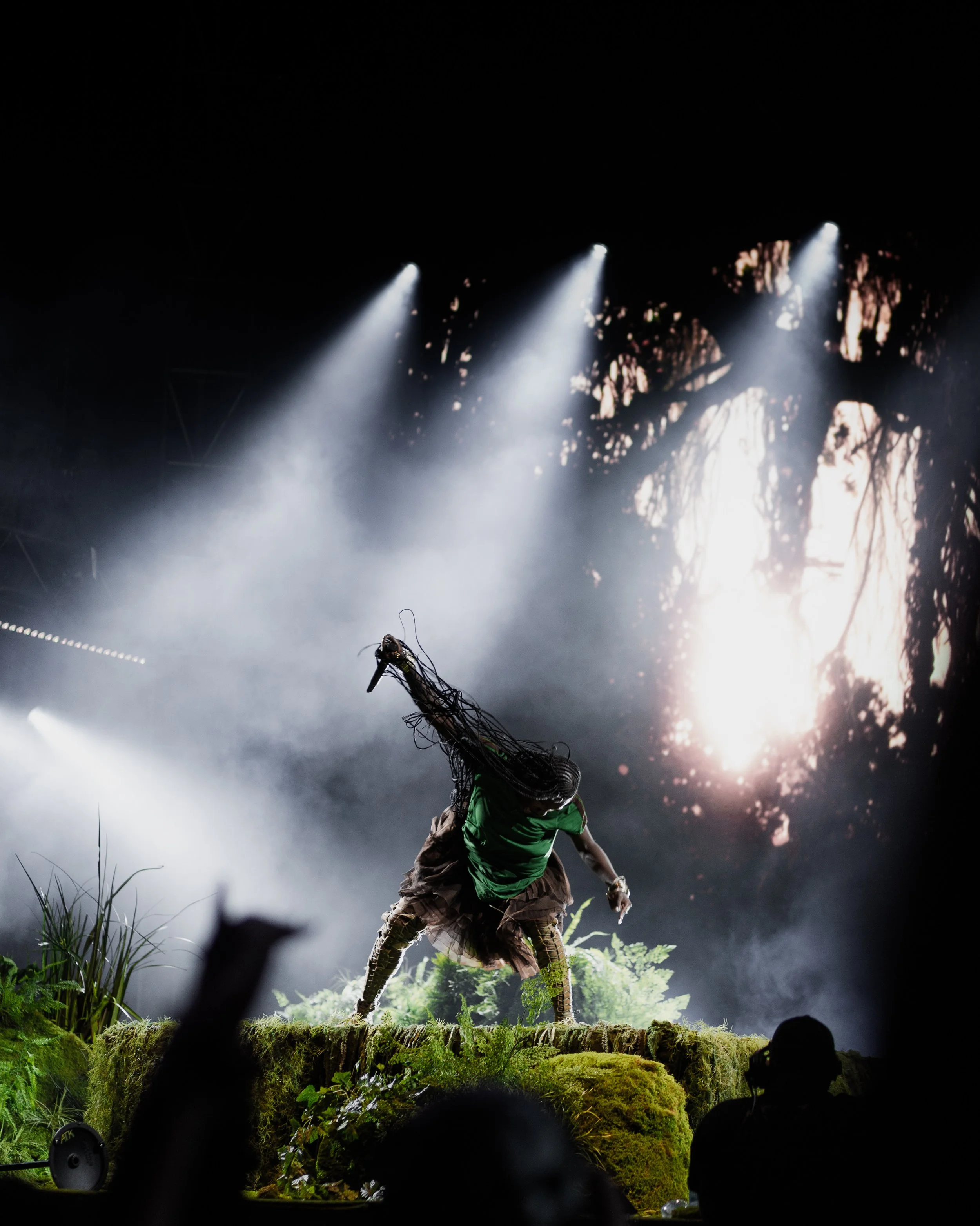Performer with long braided hair dancing on mossy stage with bright stage lights and forest backdrop.