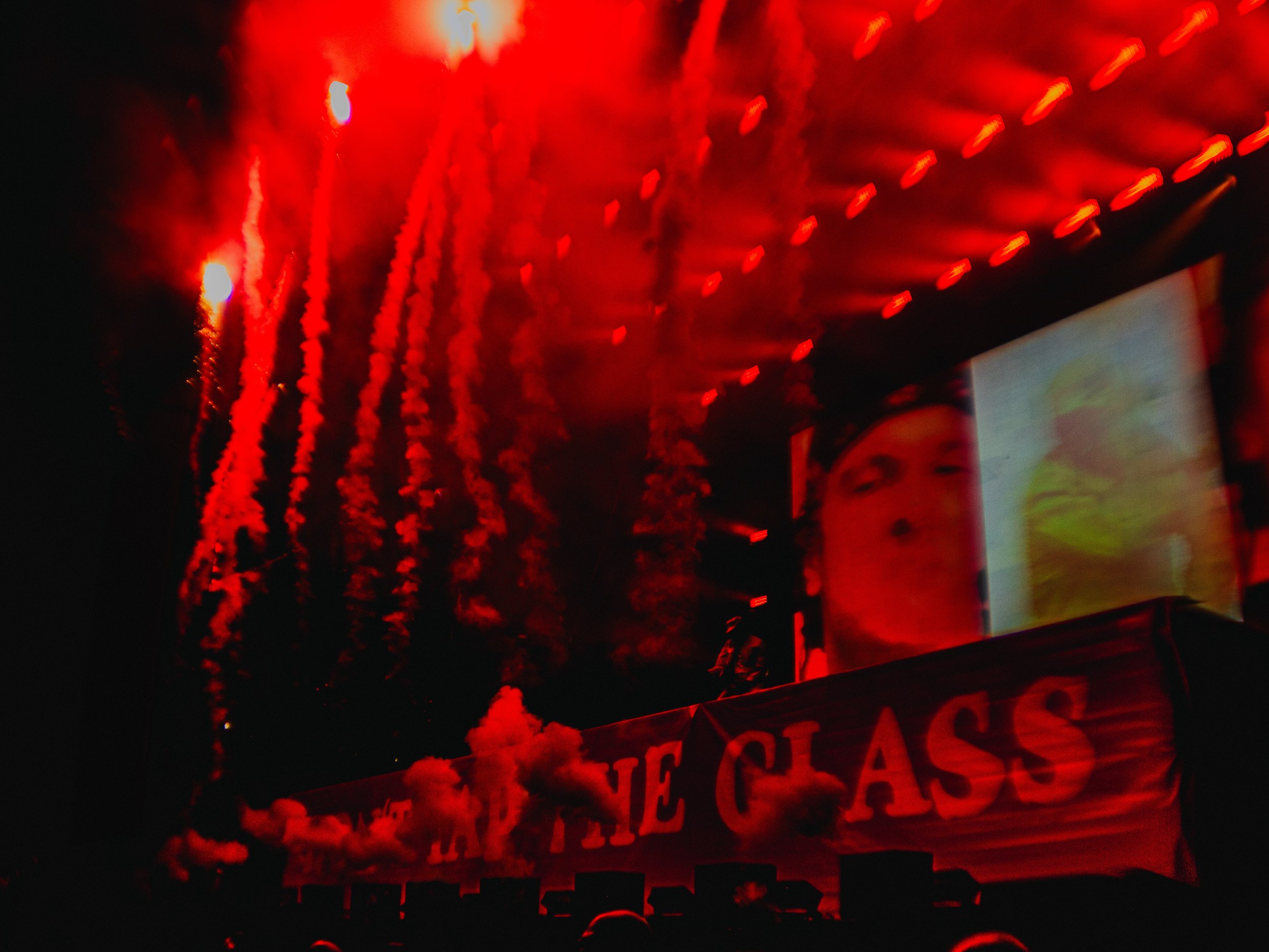 A dark scene with red fireworks lighting up the sky above a concert stage. A large screen shows a person’s face, and red streamers and smoke are visible. A banner reading 'THE CLASS' is partially visible at the bottom.