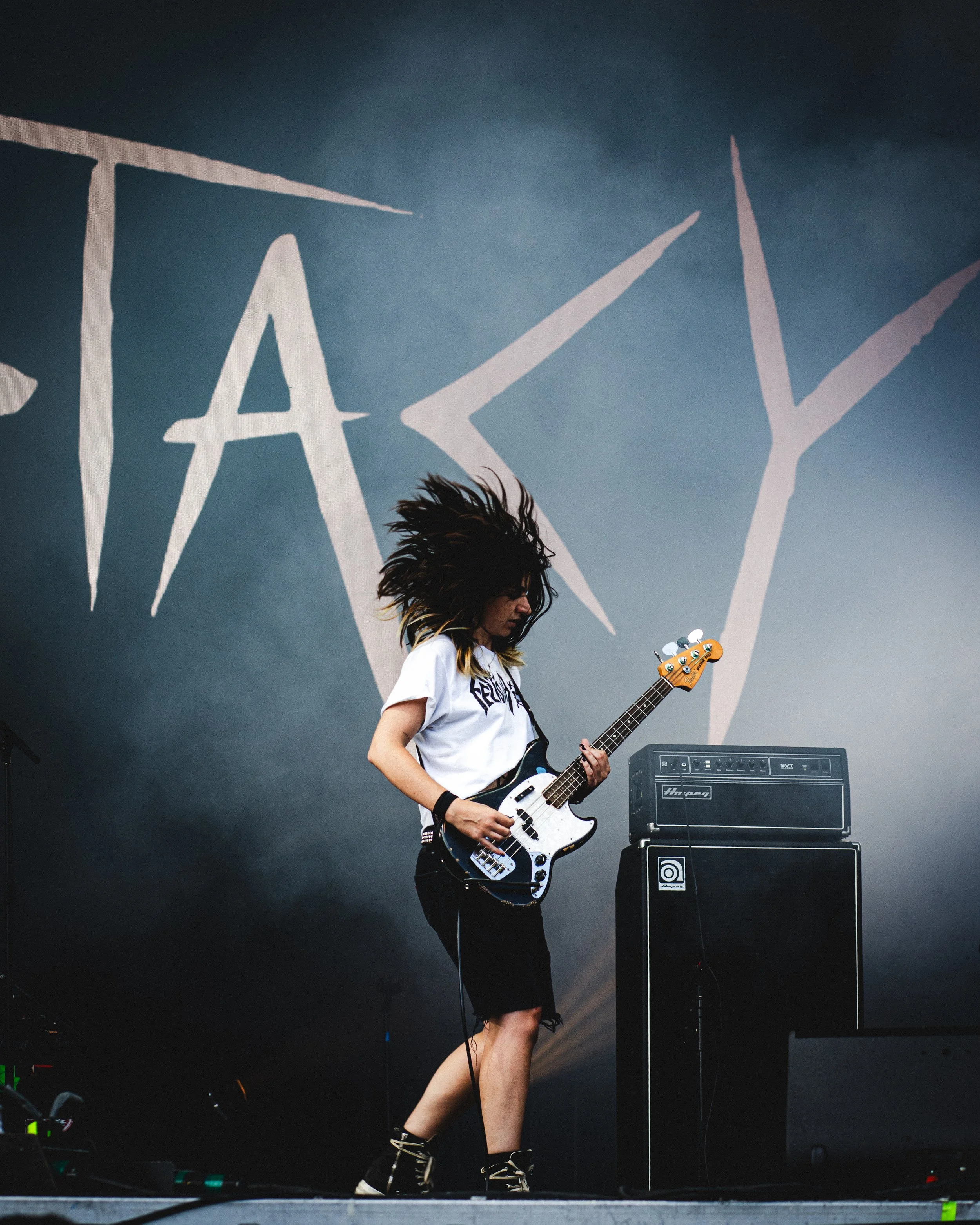 A woman with long, spiky hair playing an electric guitar on stage during a concert, with a large background sign that reads 'TAKE'