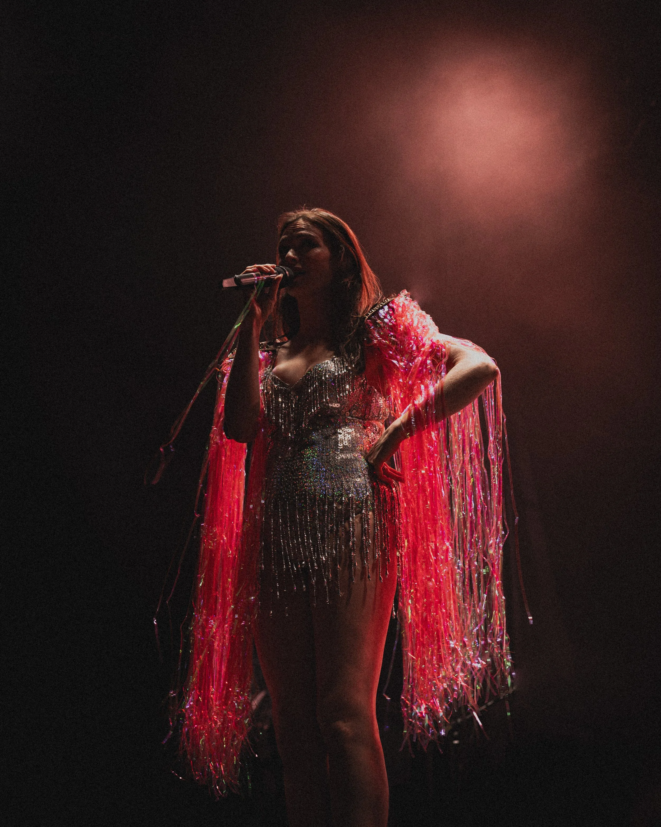 A female performer on stage singing into a microphone, wearing a glittering dress and a long, fringed pink cape, with stage lighting creating a dramatic effect.