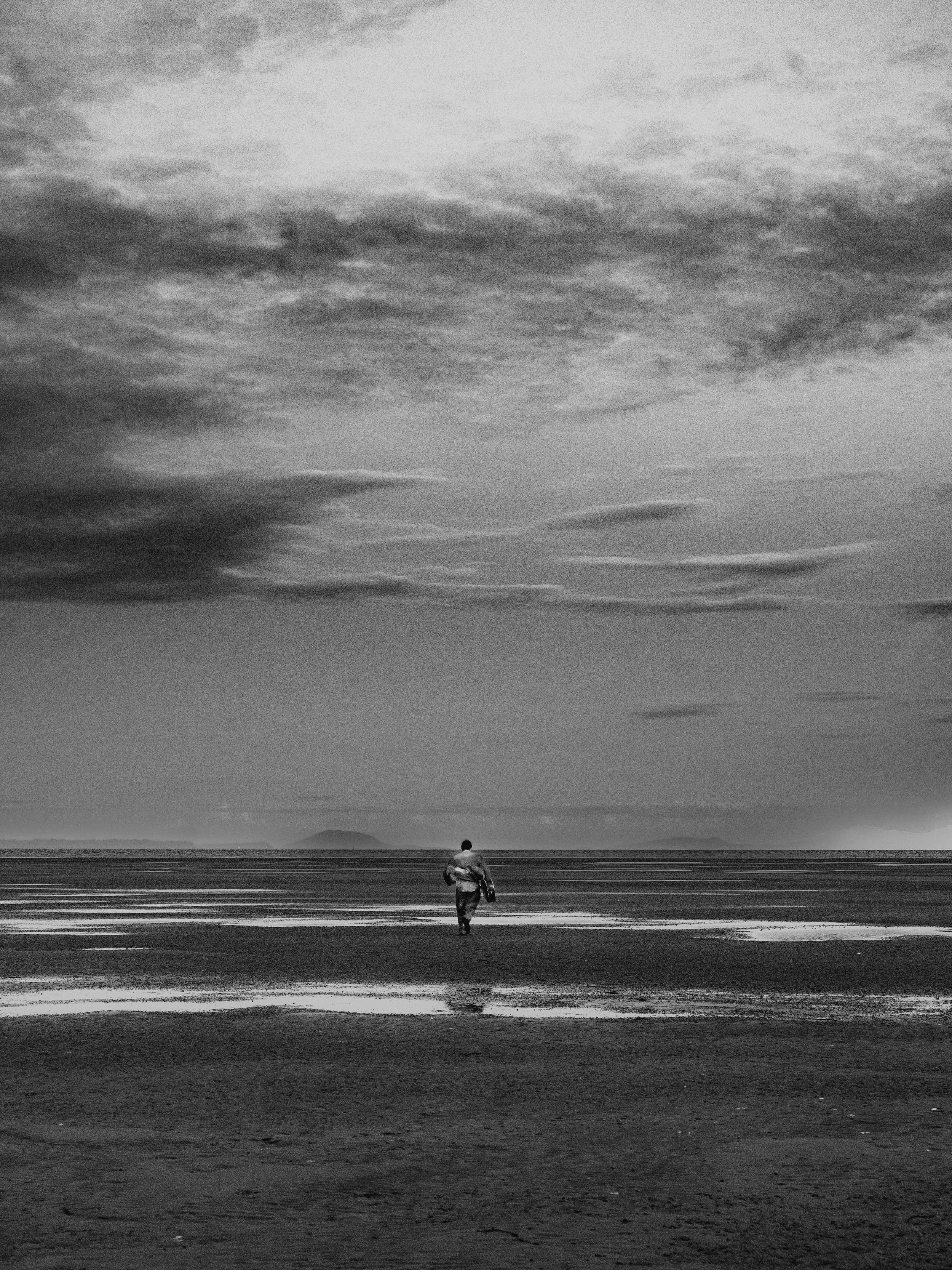 A solitary person walking on a beach at sunset, with cloudy sky overhead, black and white photograph.