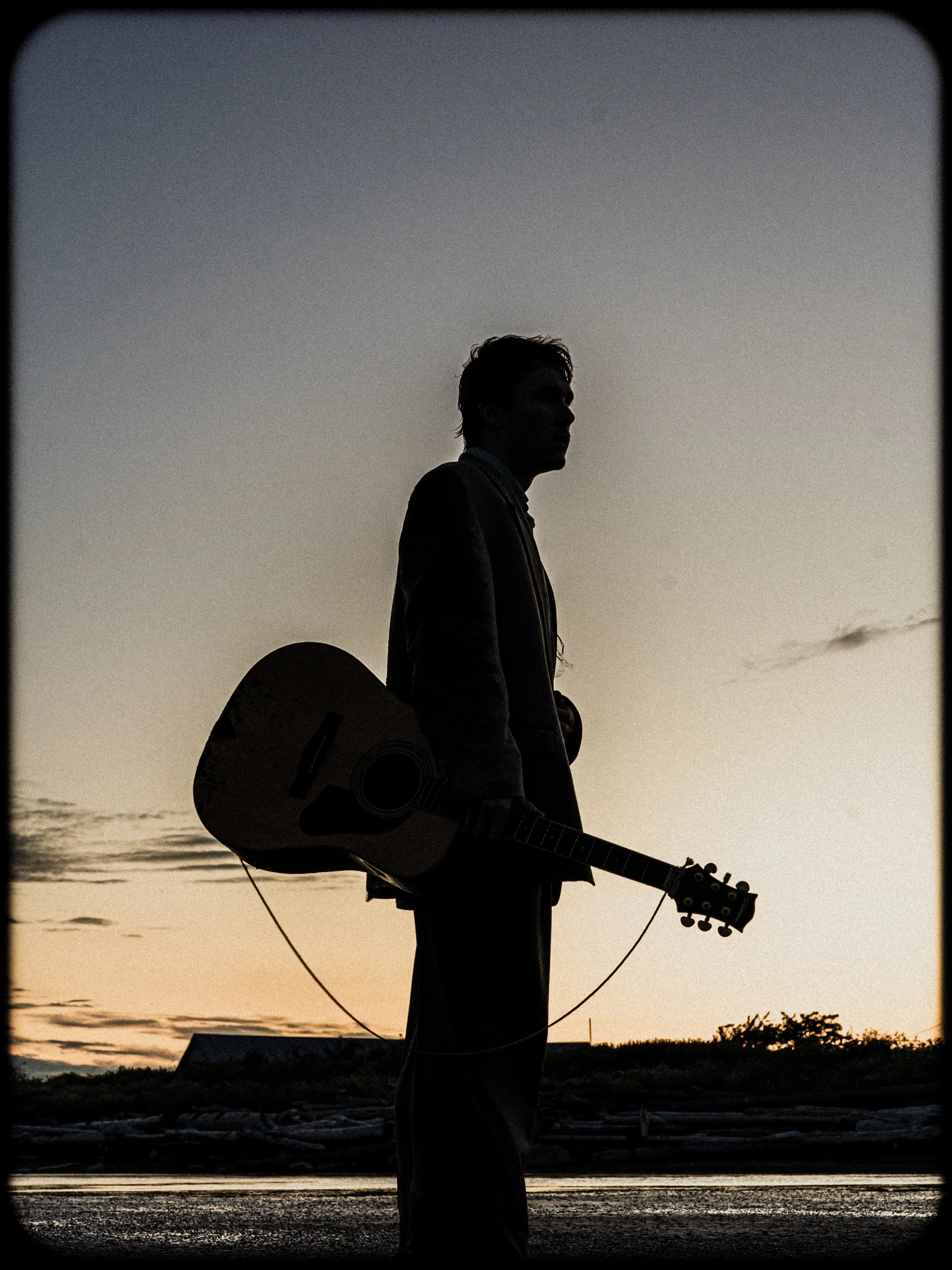 A silhouette of a person holding an acoustic guitar, standing outdoors at sunset or sunrise, with a distant horizon and a mostly clear sky.