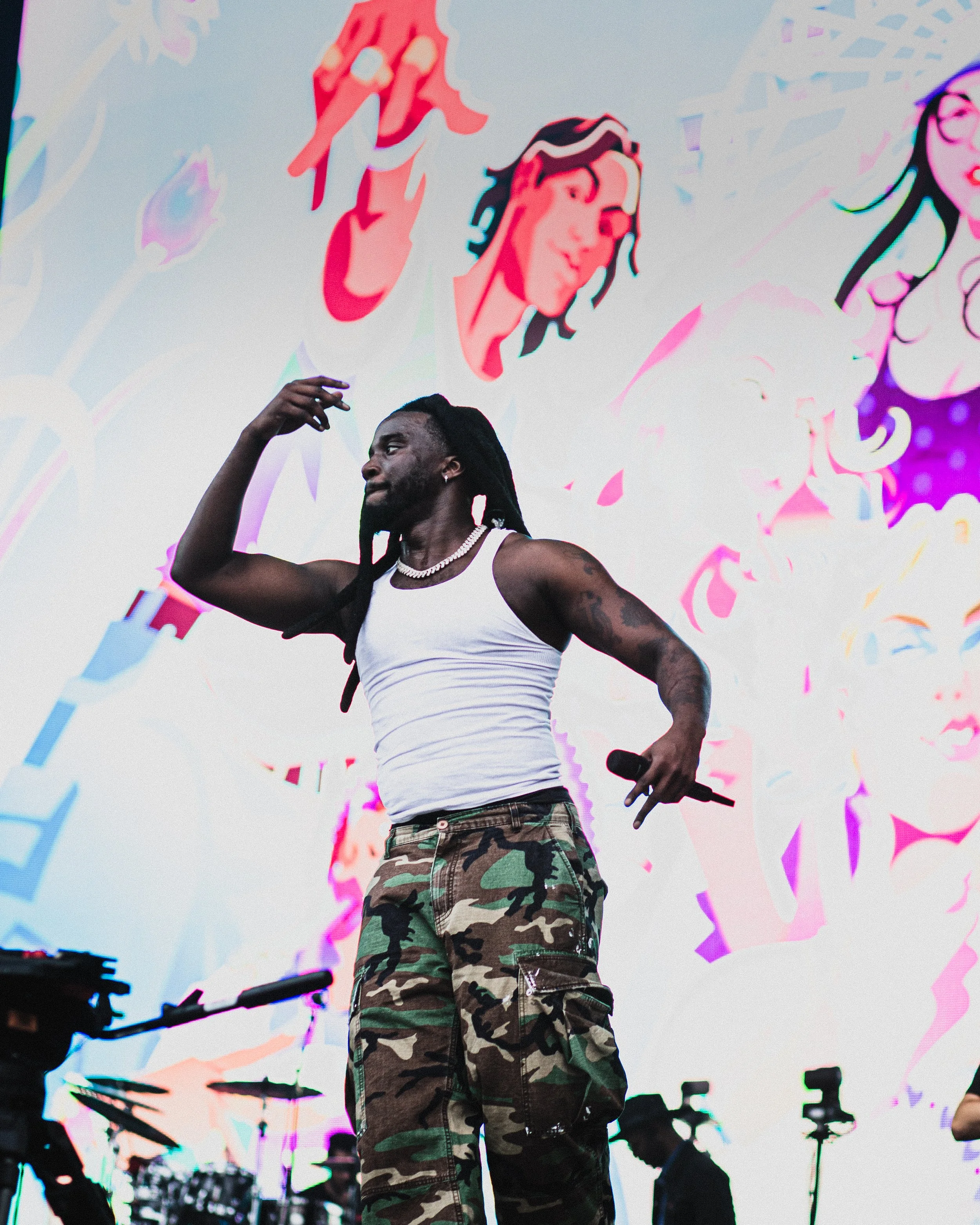 A male performer with dreadlocks, wearing a white tank top, camouflage pants, and a necklace, is holding a microphone and dancing on stage. A colorful background screen displays stylized illustrations of women with flowing hair and flowers.