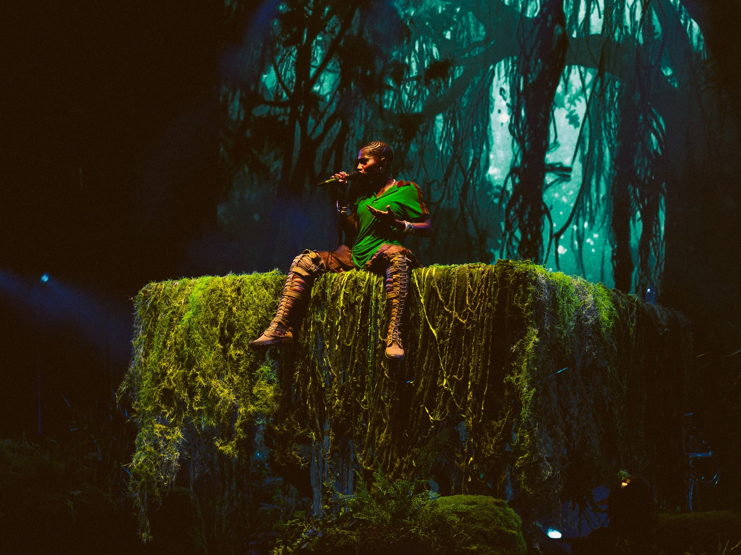 Performer sitting on a mossy platform in a forest-themed stage, holding a microphone, with a backdrop of dark, twisted trees and vines.