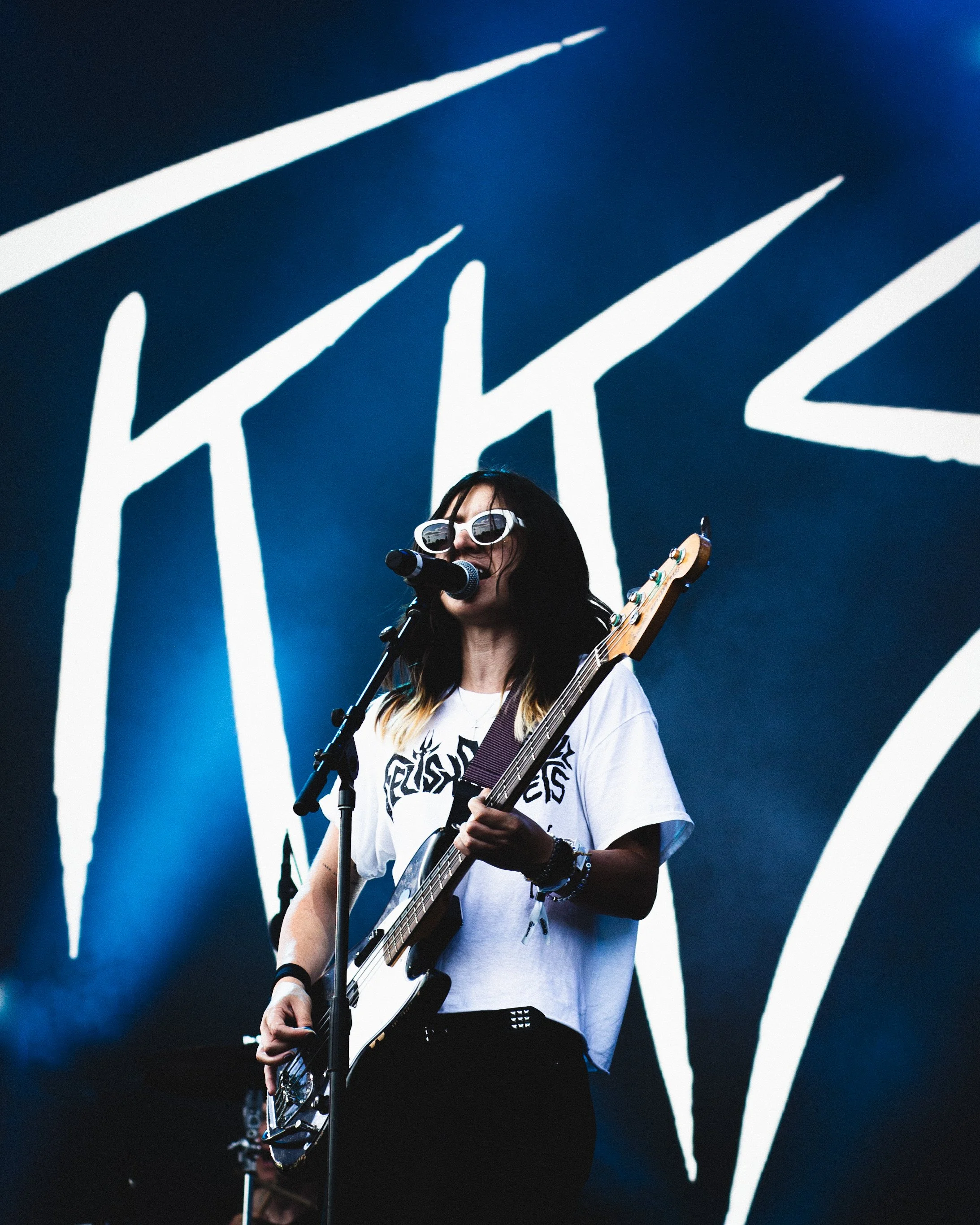 Female musician with sunglasses singing into a microphone while playing an electric guitar on stage with a blue background and white large logo or graffiti.