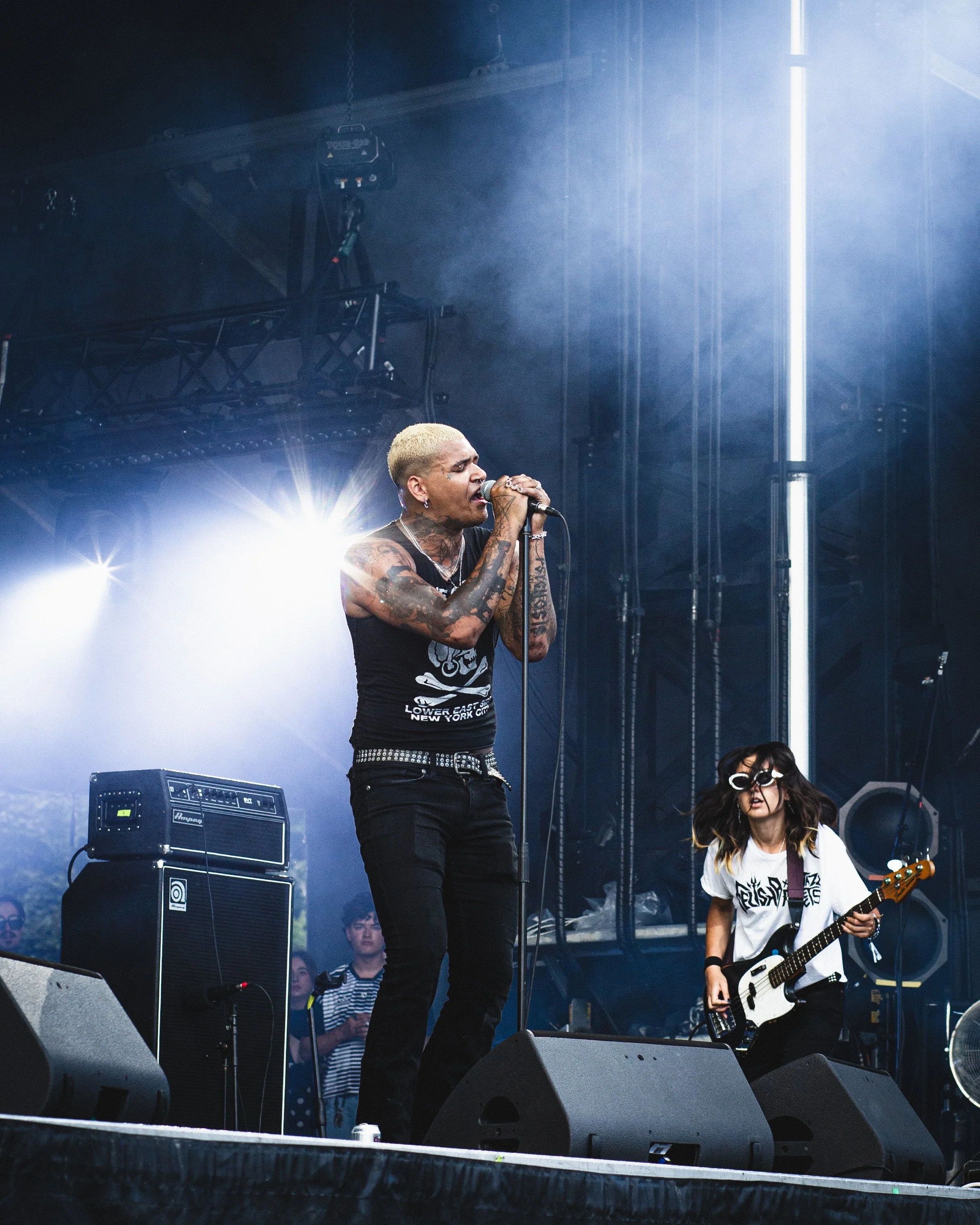 A band performs on stage at an outdoor concert, with a blonde-haired vocalist singing into a microphone and a female guitarist wearing large glasses playing a bass guitar. Bright stage lights and equipment are visible.