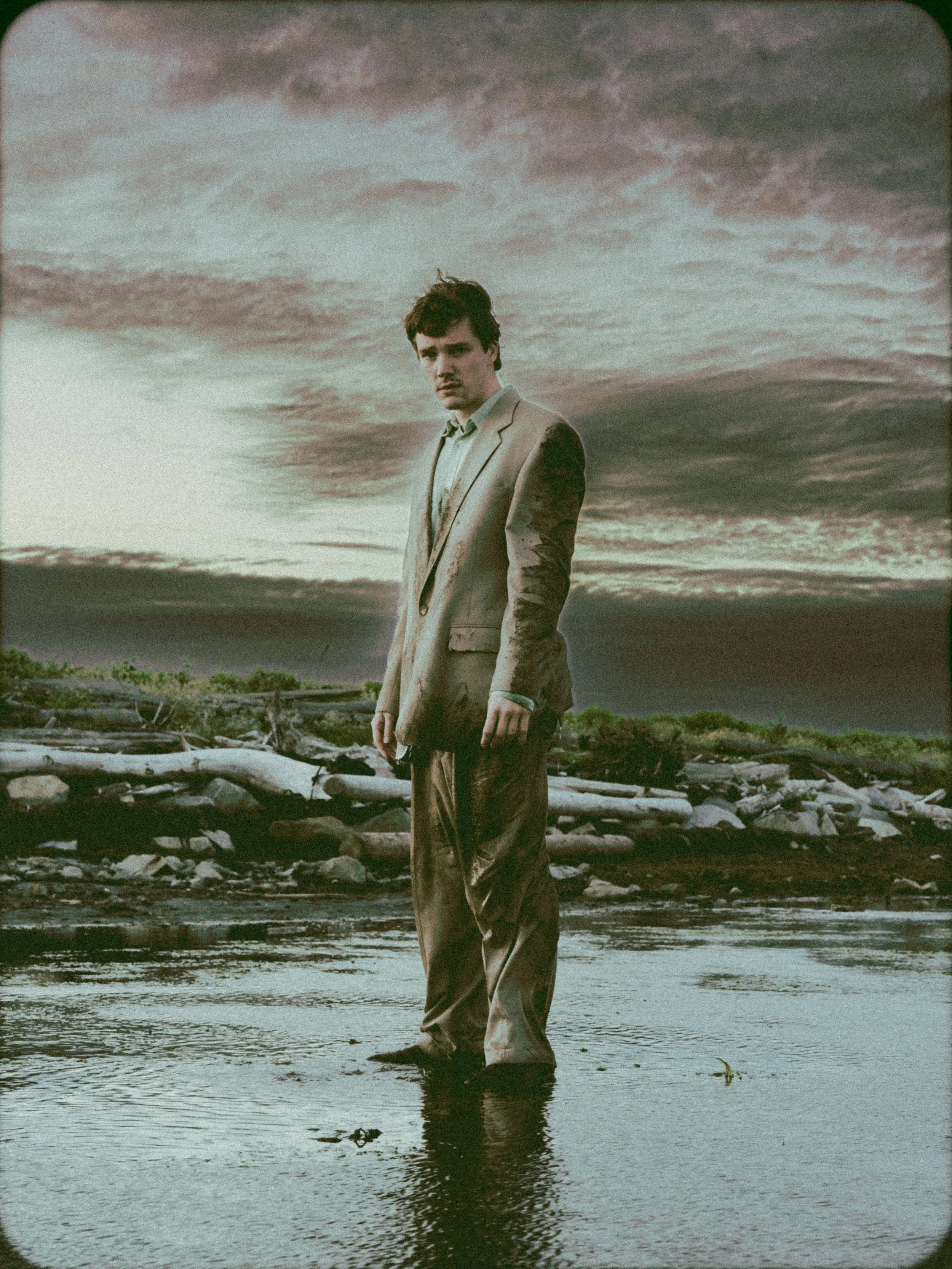 A young man in a beige suit standing in shallow water, with a river or stream, driftwood, and a cloudy, stormy sky in the background.