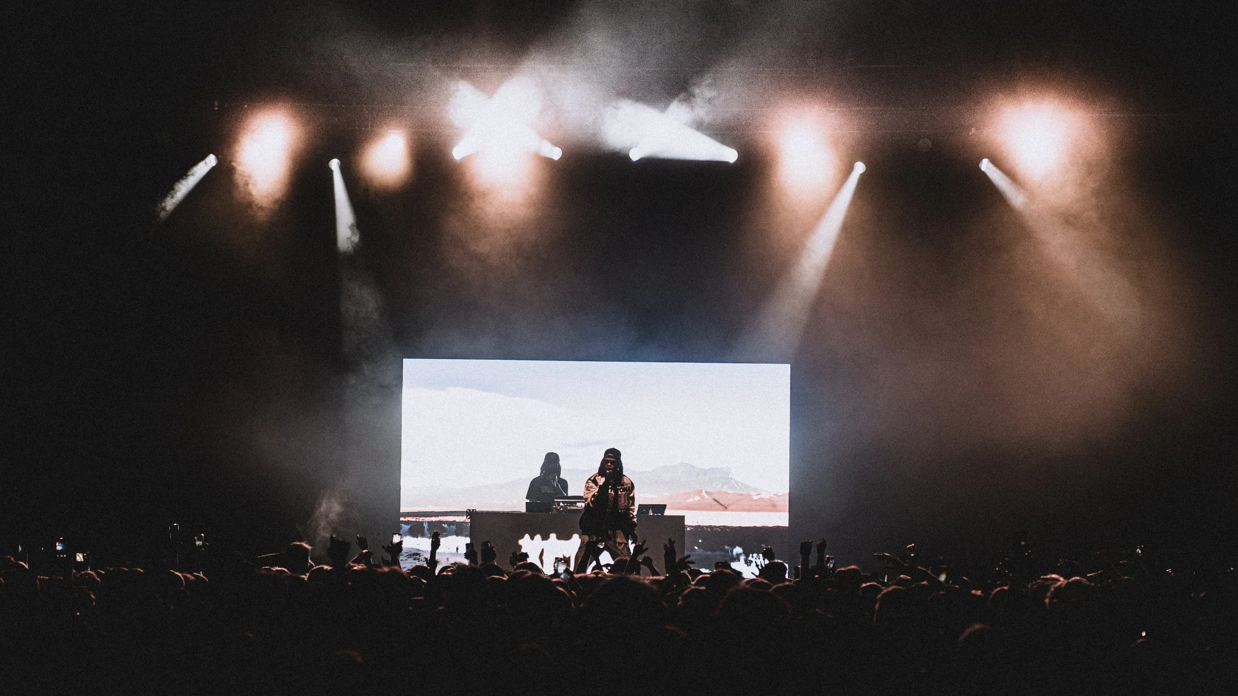 A live concert with a performer on stage holding a microphone, backlit against a large screen displaying a desert landscape. The stage is illuminated with several bright spotlights, and the audience in the foreground has many hands raised, some holdi