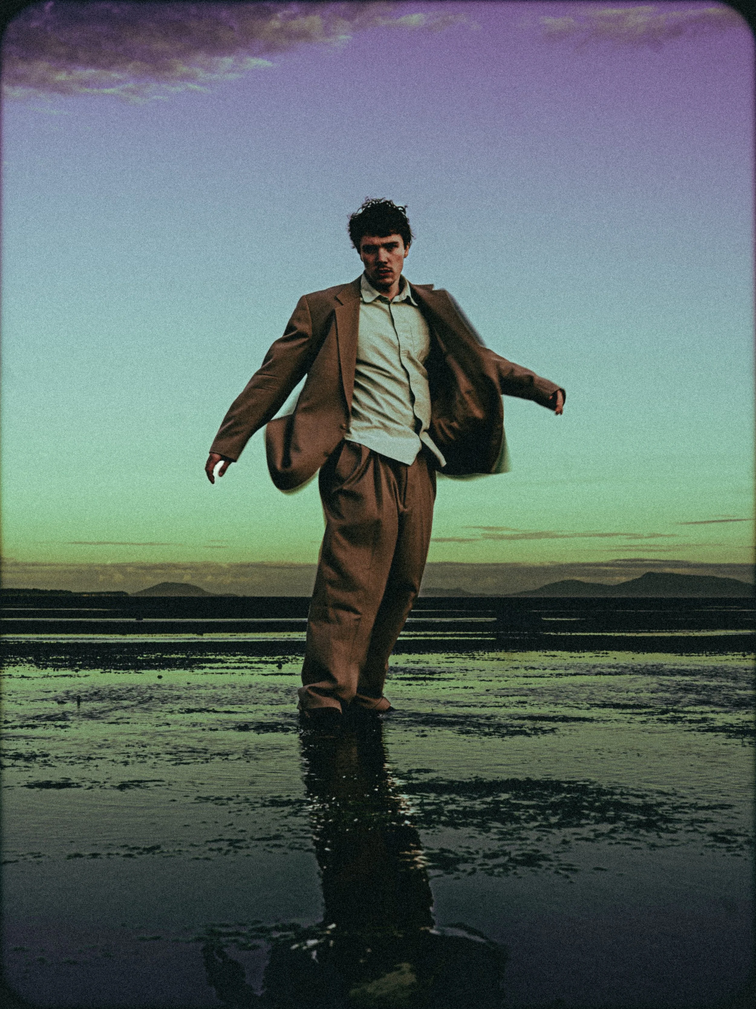 A man in a brown suit stands in shallow water during a sunset or sunrise, with a sky filled with soft purple and orange clouds and distant mountains in the background.