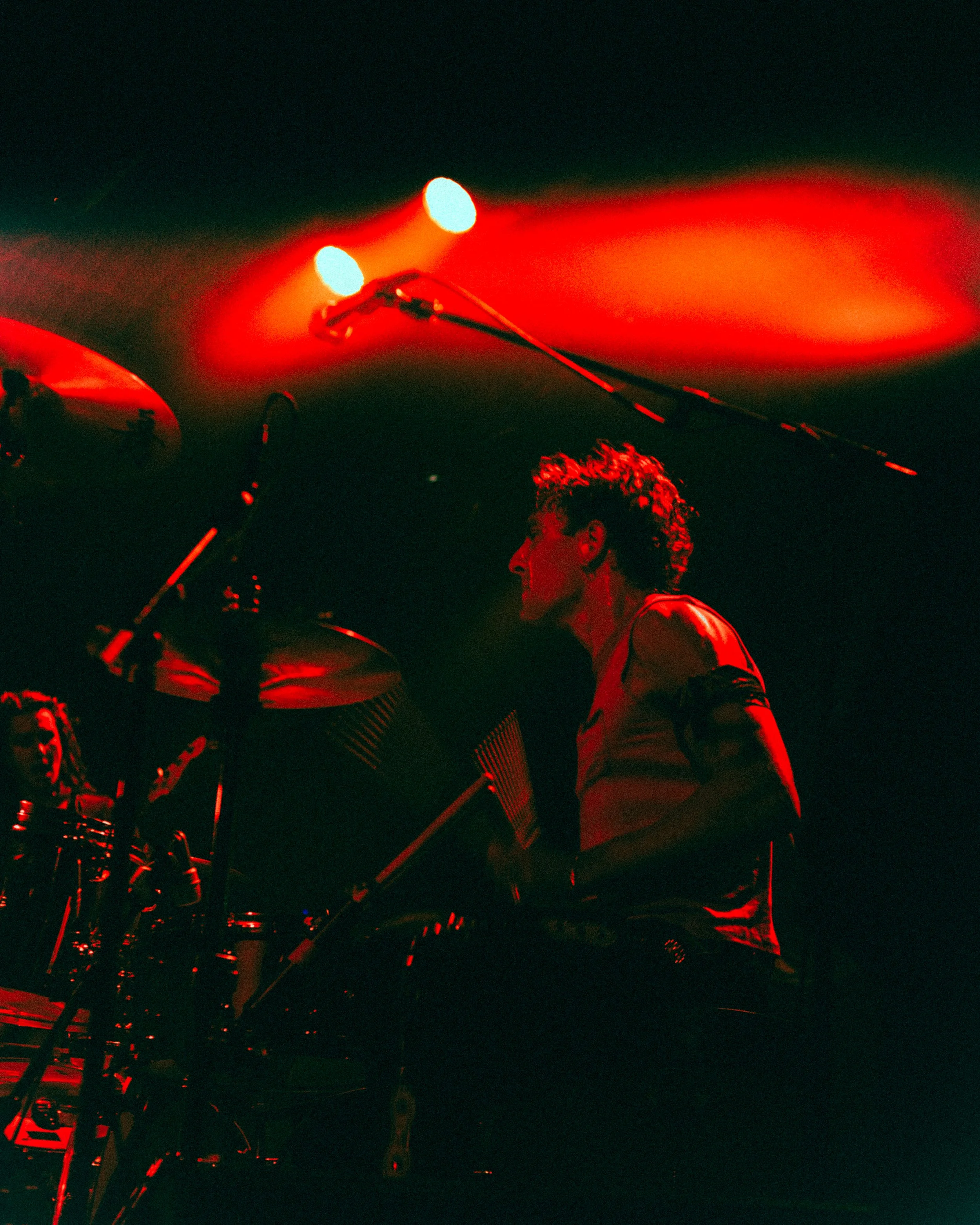 A musician with curly hair playing drums on stage, illuminated by red stage lights with a dark background.