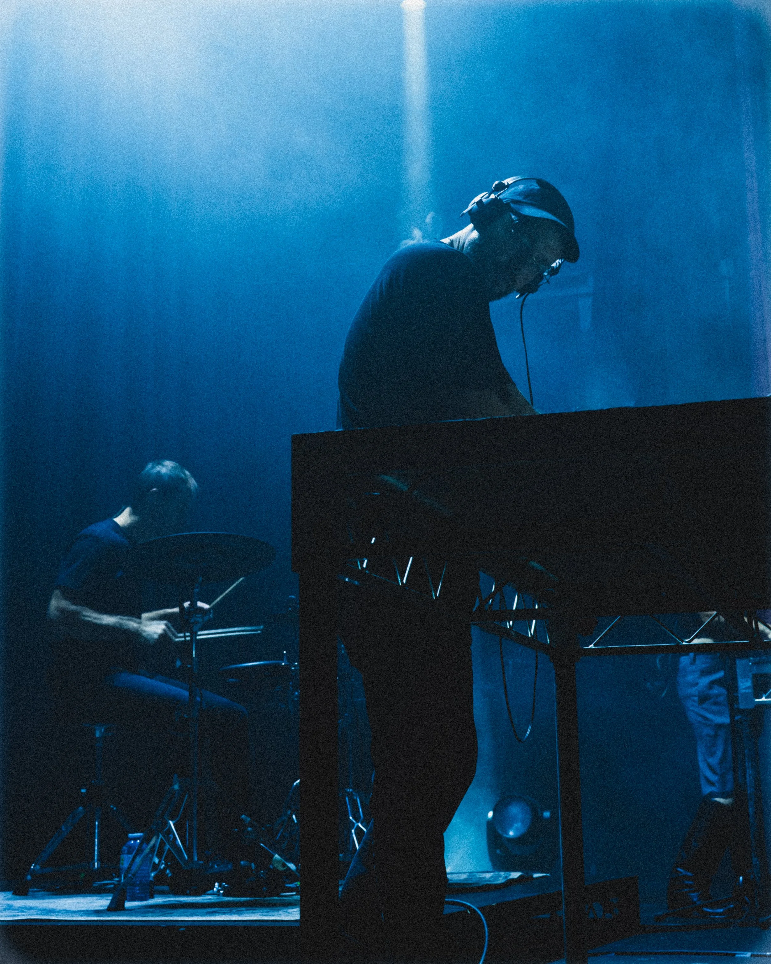 Two musicians performing on stage under blue lighting, one playing drums and the other using electronic equipment.
