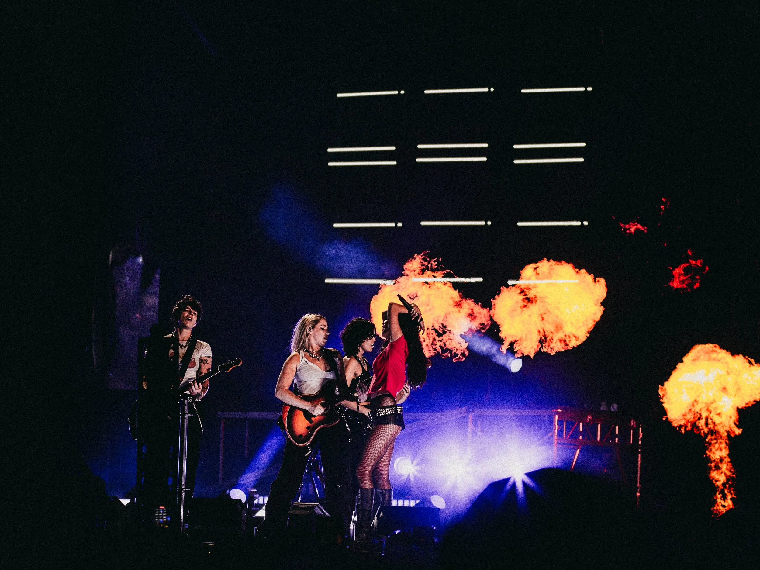 A band performing on stage with flames and fire effects behind them, and bright stage lights illuminating the scene.