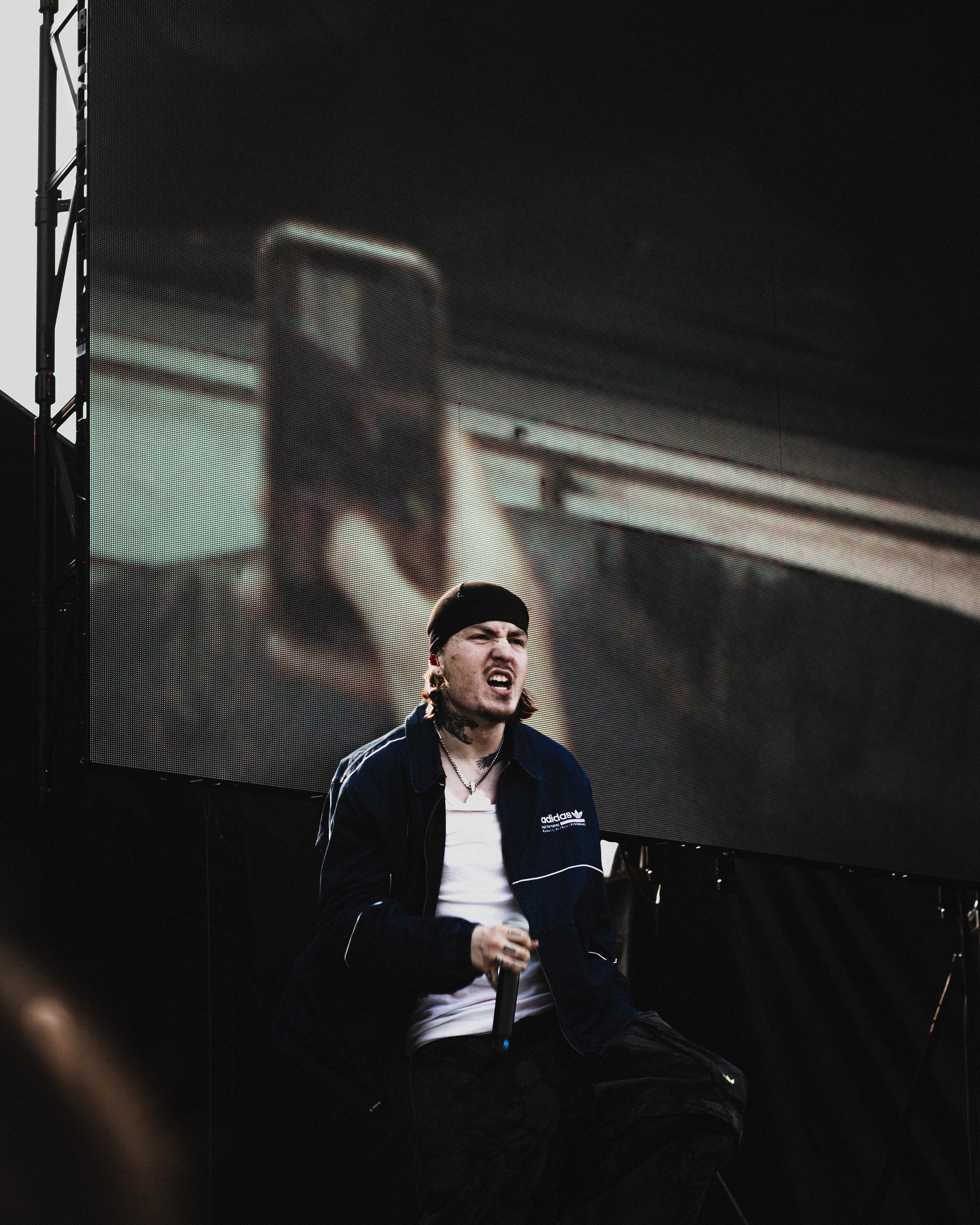 A male singer with long hair, a black beanie, and piercings, wearing a navy jacket and white shirt, singing into a microphone on stage with a large screen behind him displaying a distorted image of a person holding a cell phone.