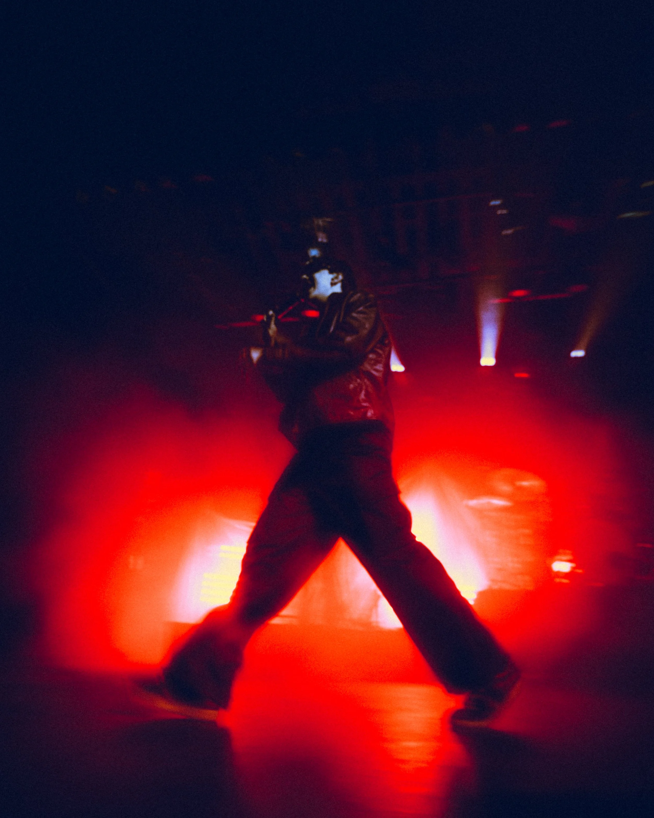A person in dark clothing singing into a microphone on a stage illuminated by red lights.