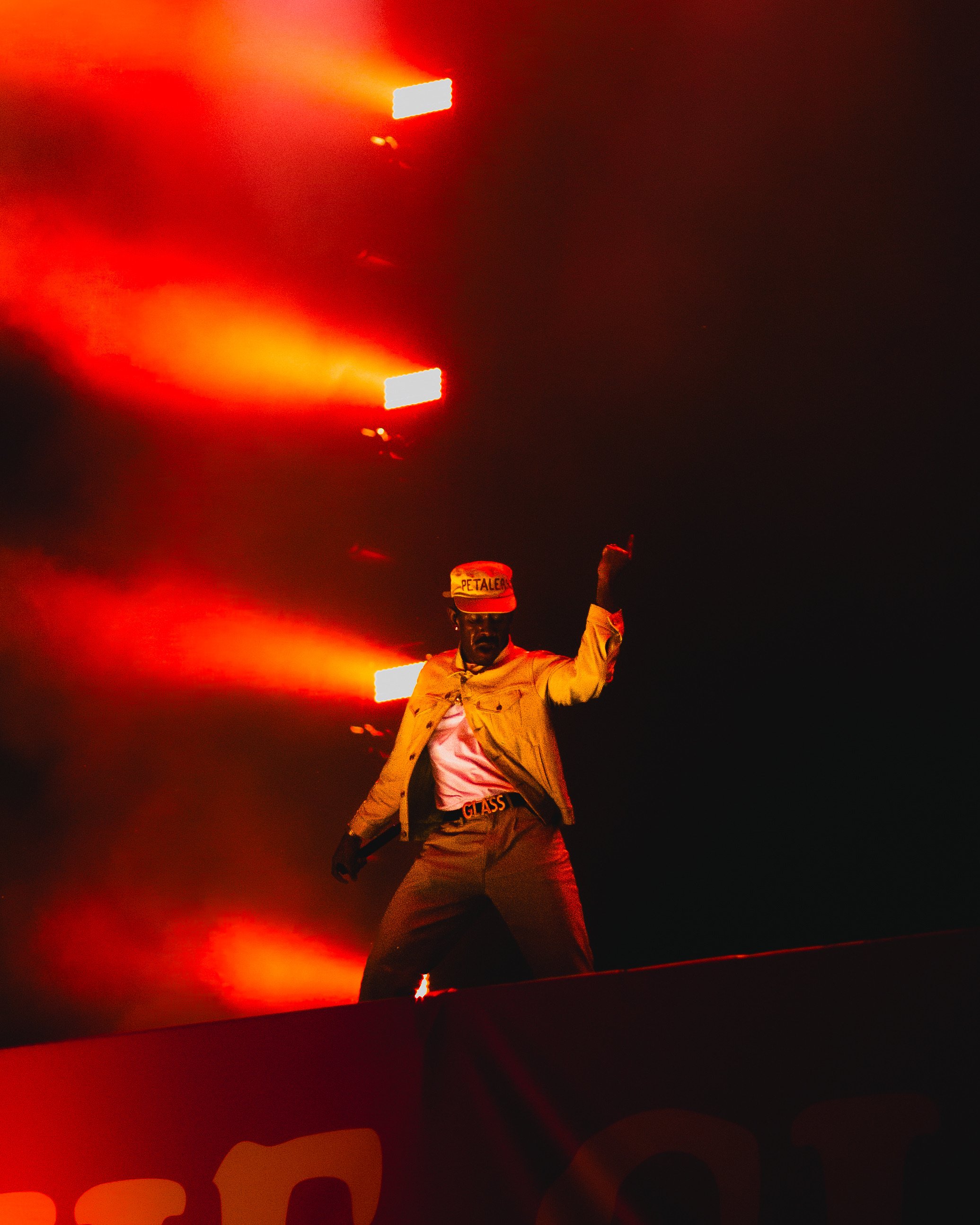 A man standing on a stage at night, illuminated by bright red lights, wearing a yellow jacket, beige pants, a white shirt, and a red cap with the words 'PETALER' on it, posing with one arm raised.