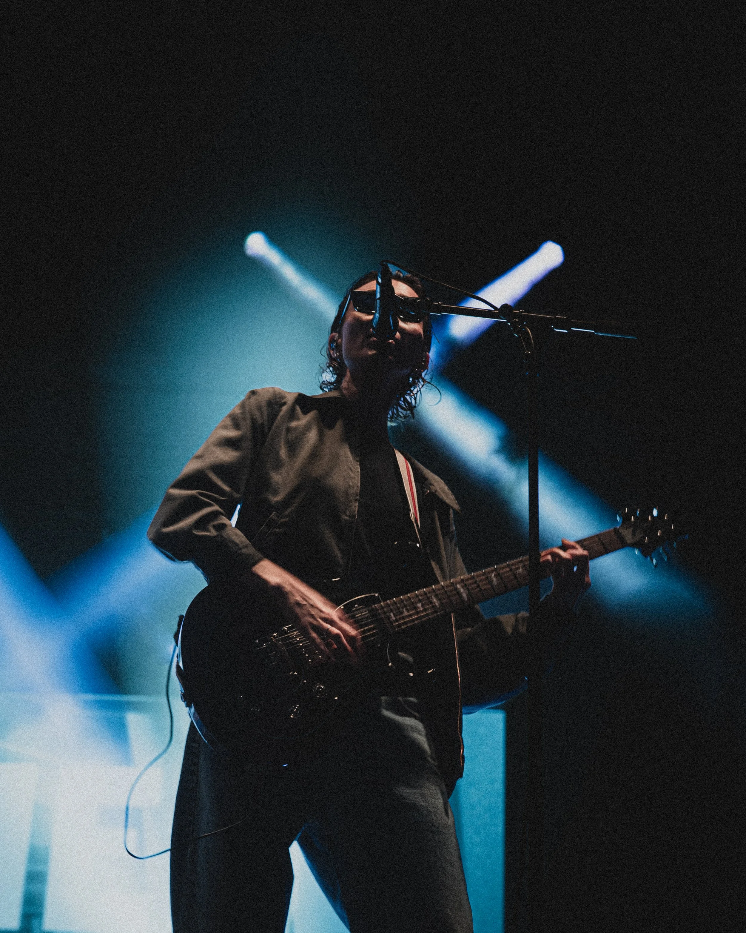 A musician performing on stage with an electric guitar, illuminated by blue and white stage lighting, wearing sunglasses and a dark outfit.