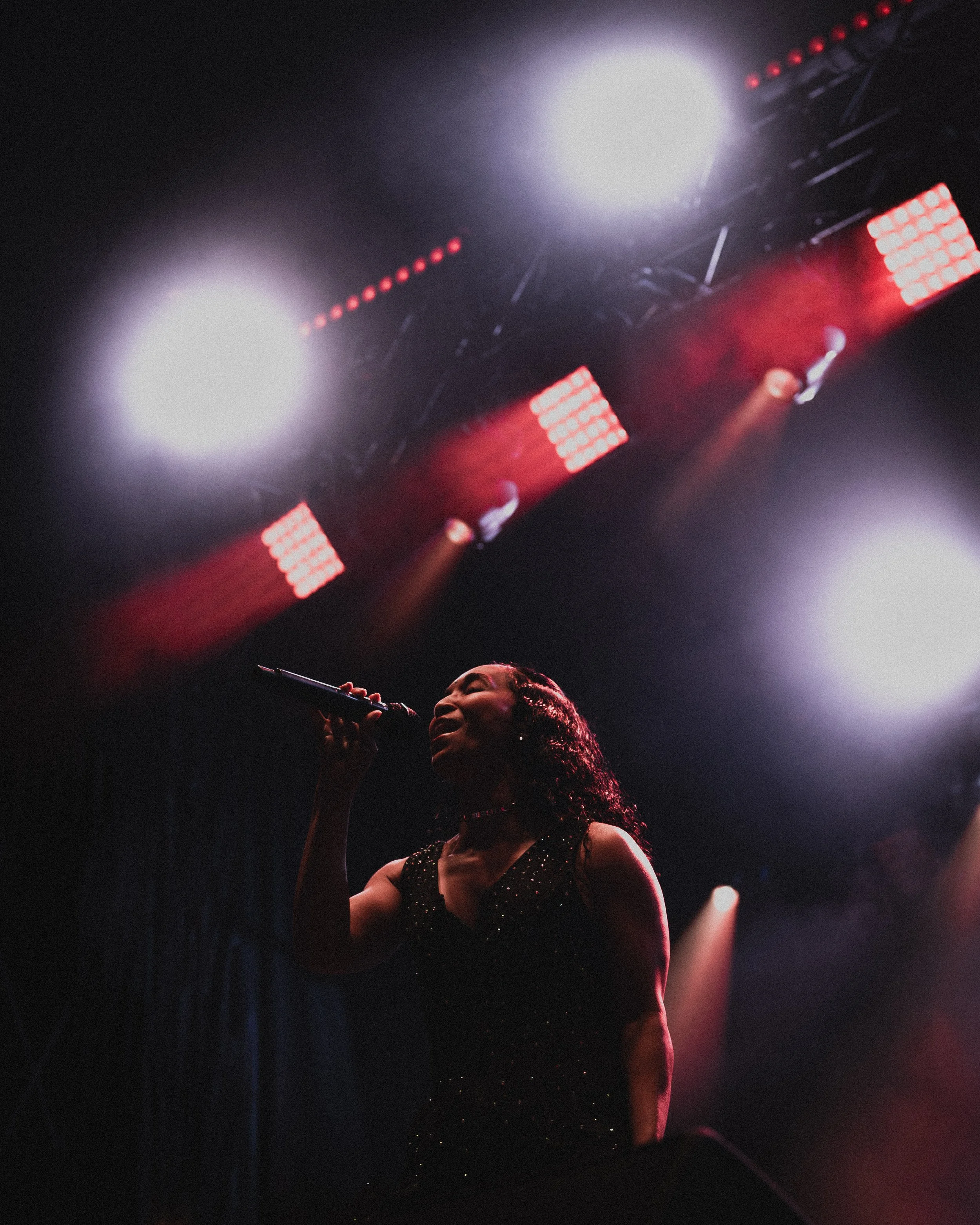 A woman singing on stage under bright overhead lights, holding a microphone, dressed in a sparkly black dress.