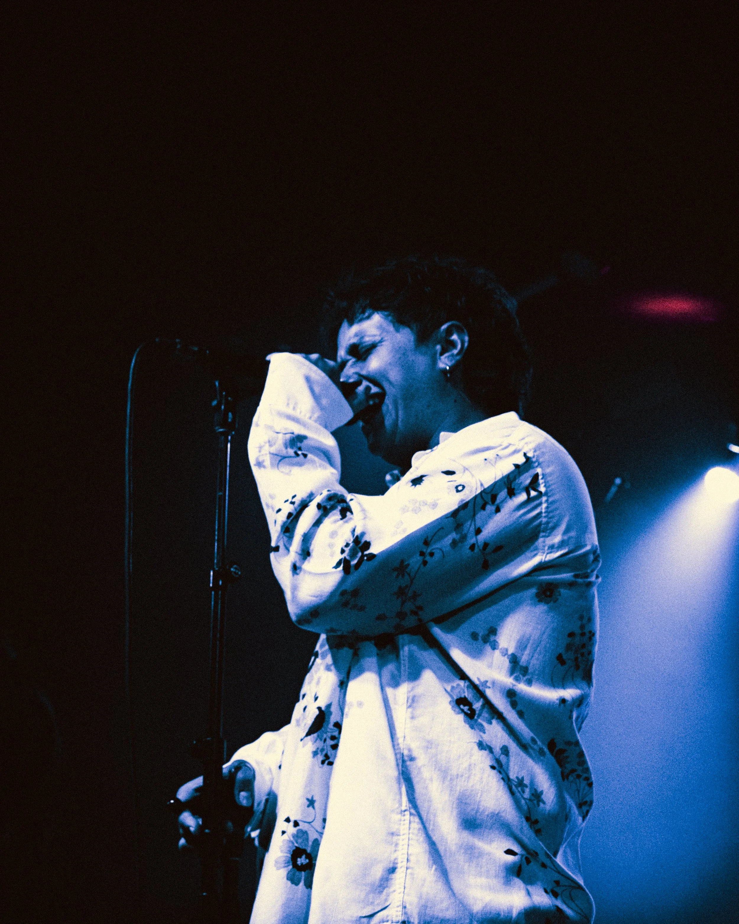 A singer performs passionately on stage, holding a microphone close to her mouth, wearing a white floral jacket with dark floral patterns, under blue stage lighting.