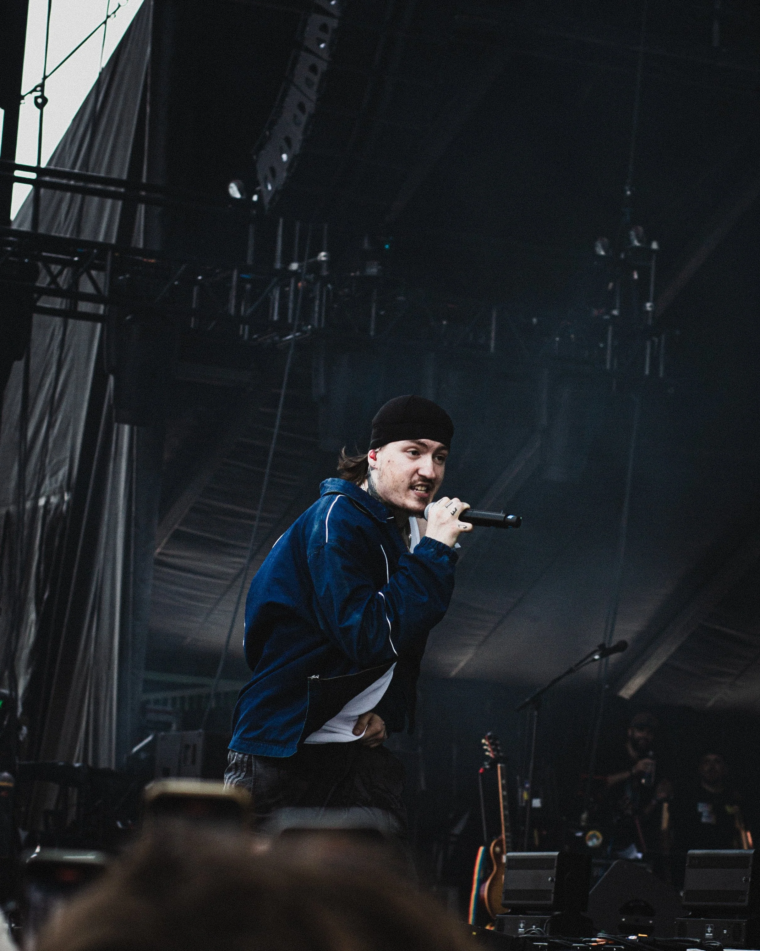 A male performer wearing a black beanie and a blue jacket is singing into a microphone on stage during a concert, with musical equipment and stage lighting visible in the background.