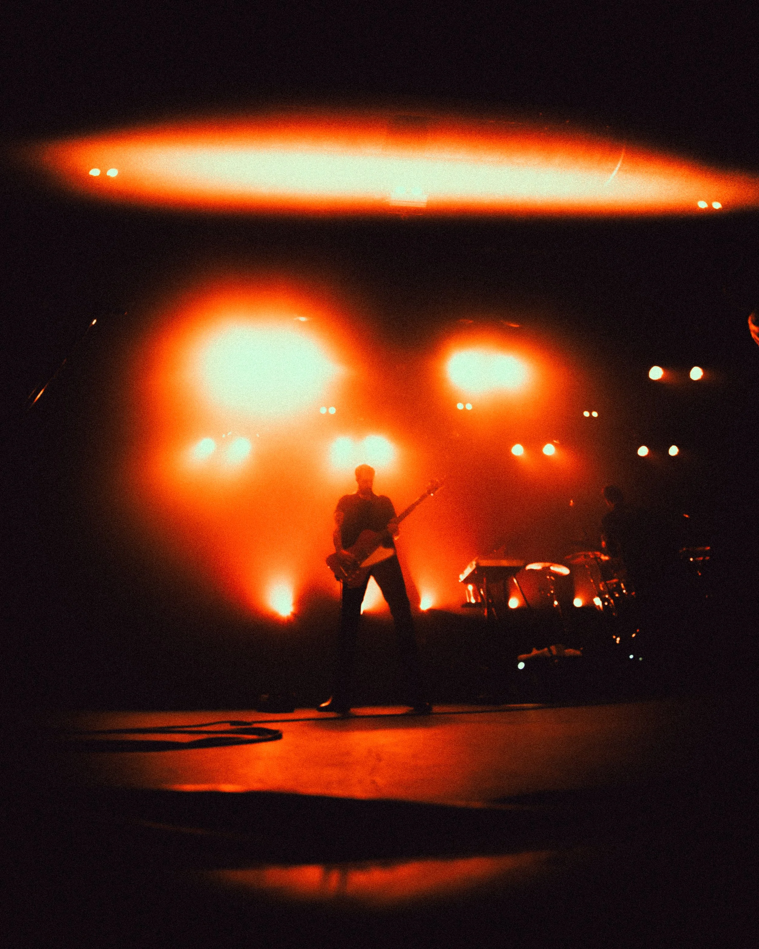 A live music performance featuring a silhouetted guitarist on stage illuminated by orange-red lights with other band members and instruments visible.