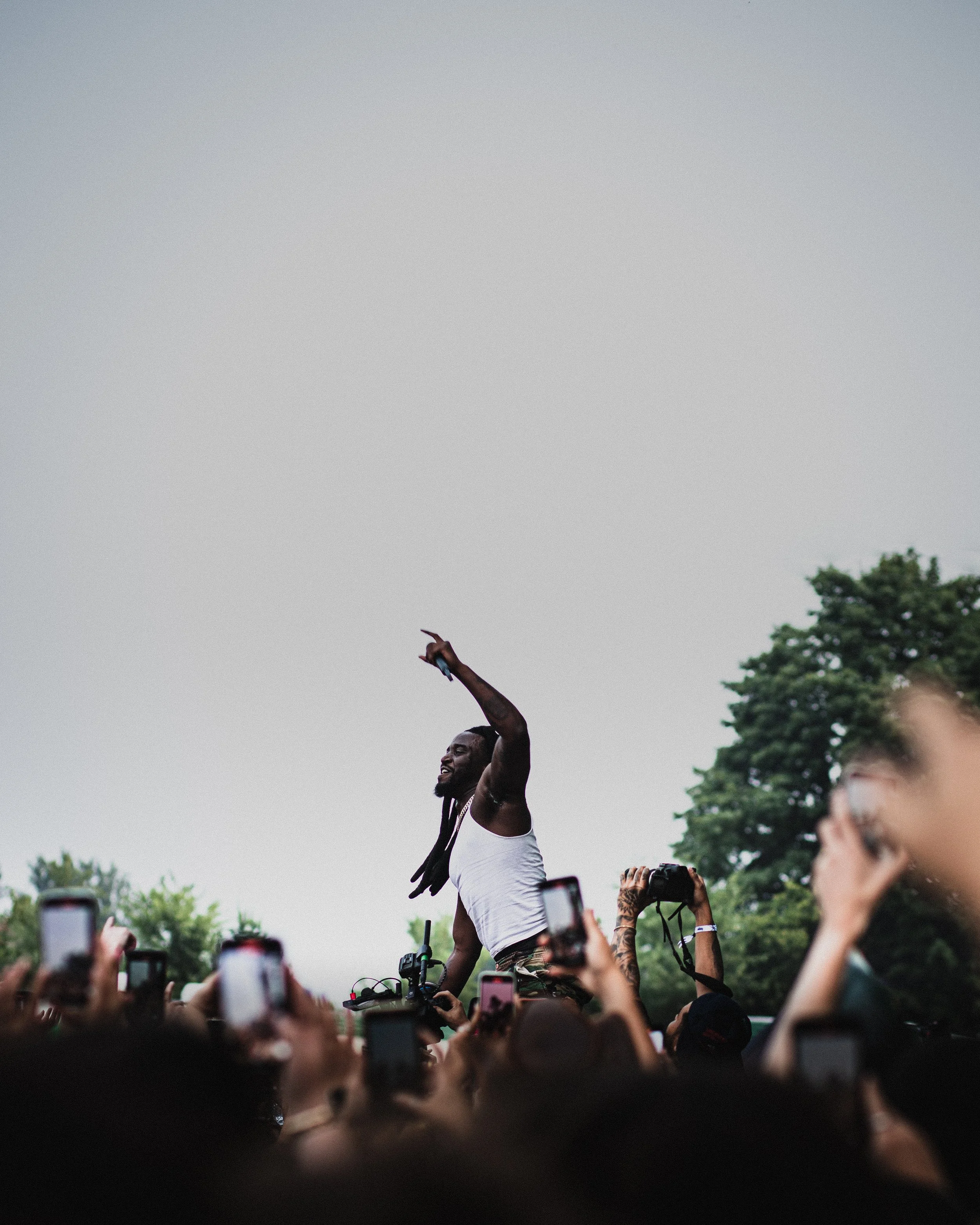 A male performer with dreadlocks and a white tank top on stage at an outdoor concert, surrounded by audience members taking photos.