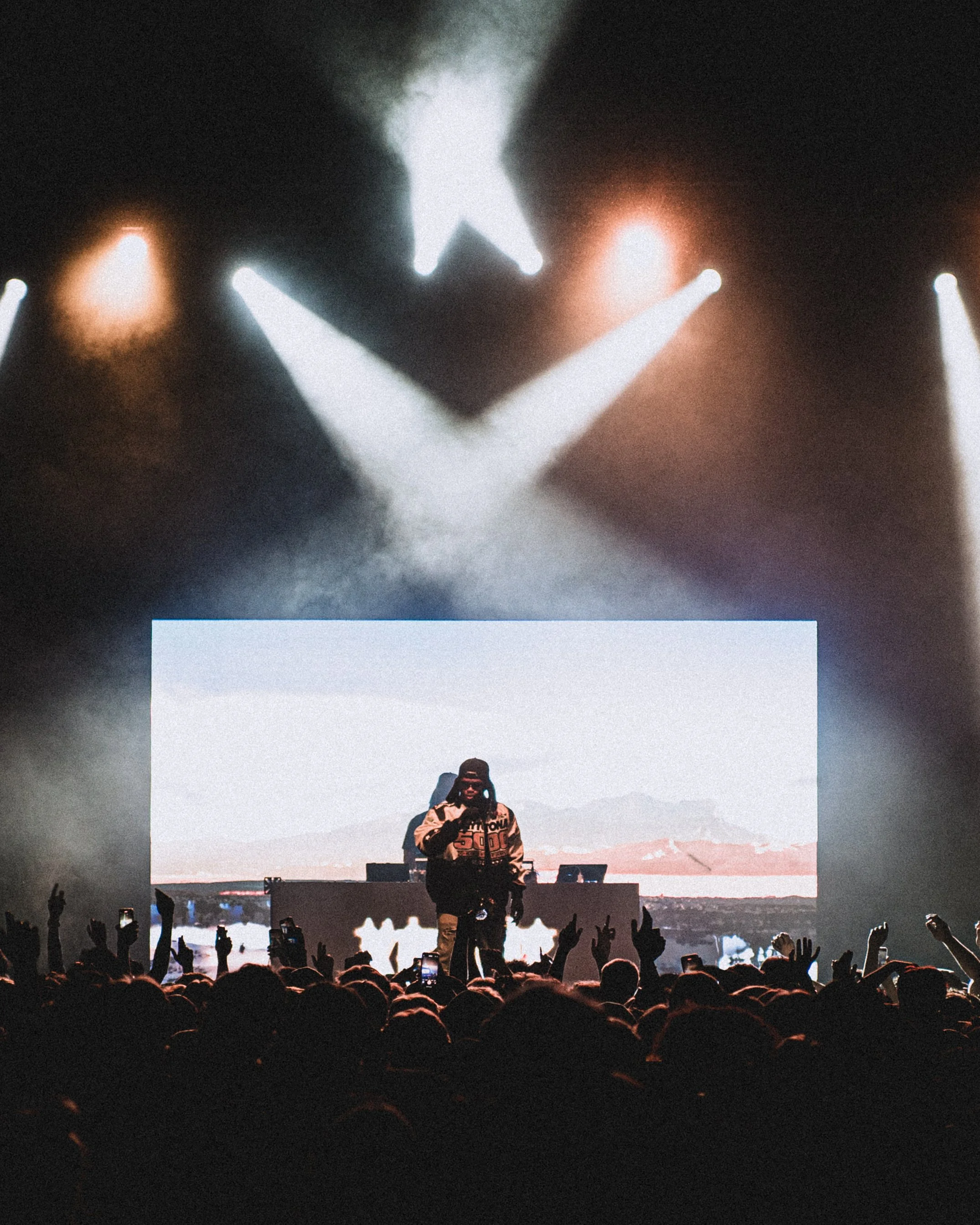 A concert scene with bright stage lights overhead, a performer on stage in front of a large digital screen, audience members taking photos, some with raised hands.