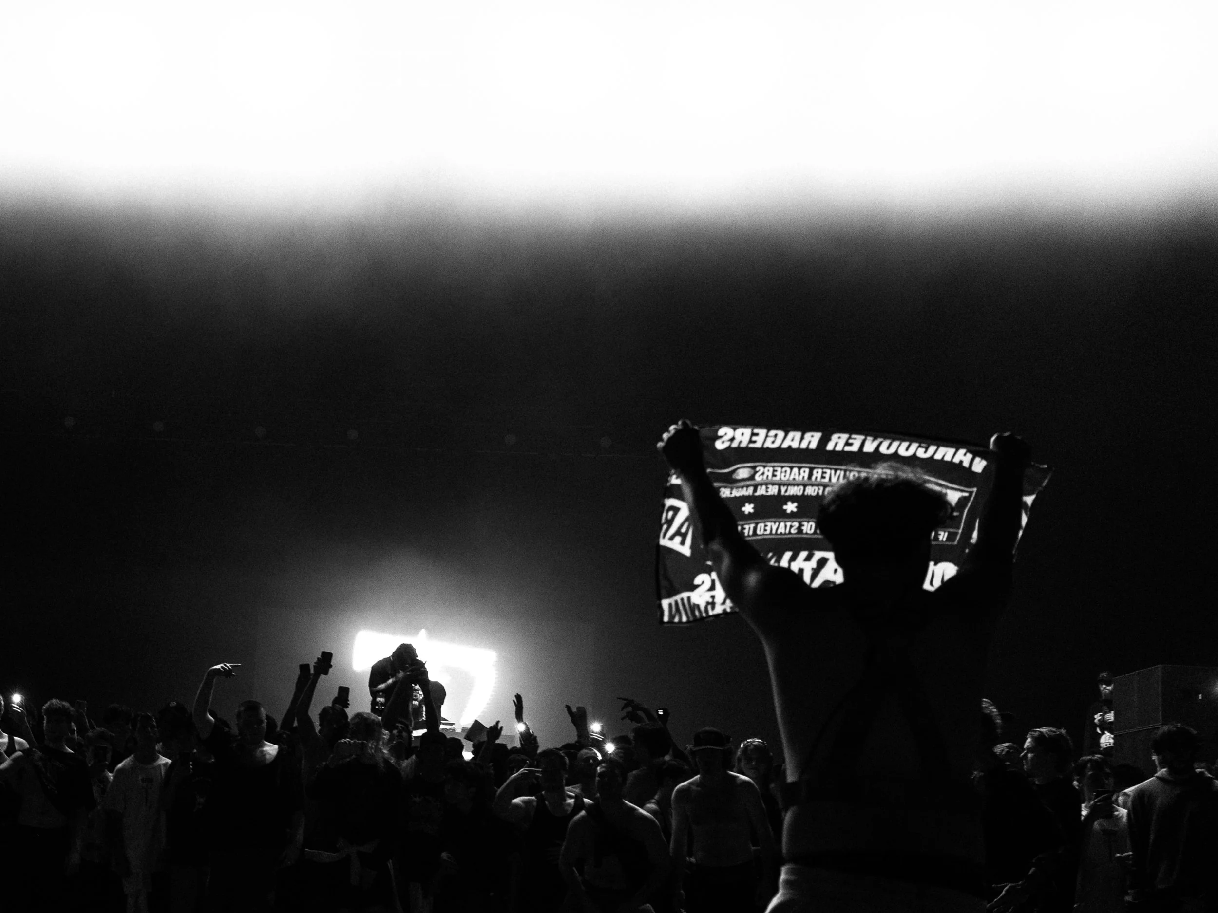 A concert scene in black and white with a large crowd. A person in the foreground is holding a flag with text, and a performer is on stage with bright backlighting.