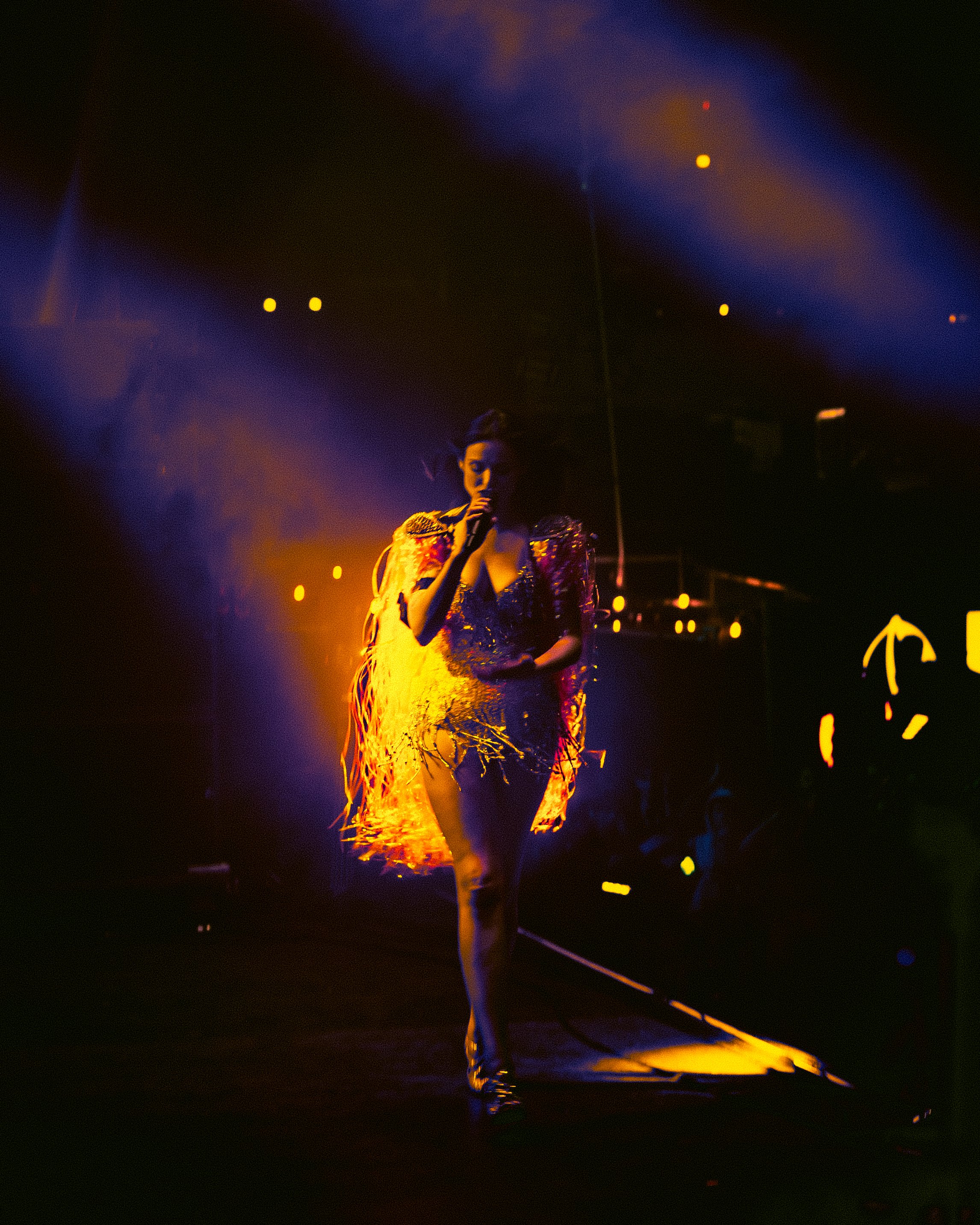 A woman walking on stage during a performance, holding a microphone, with dramatic lighting creating a silhouette effect.