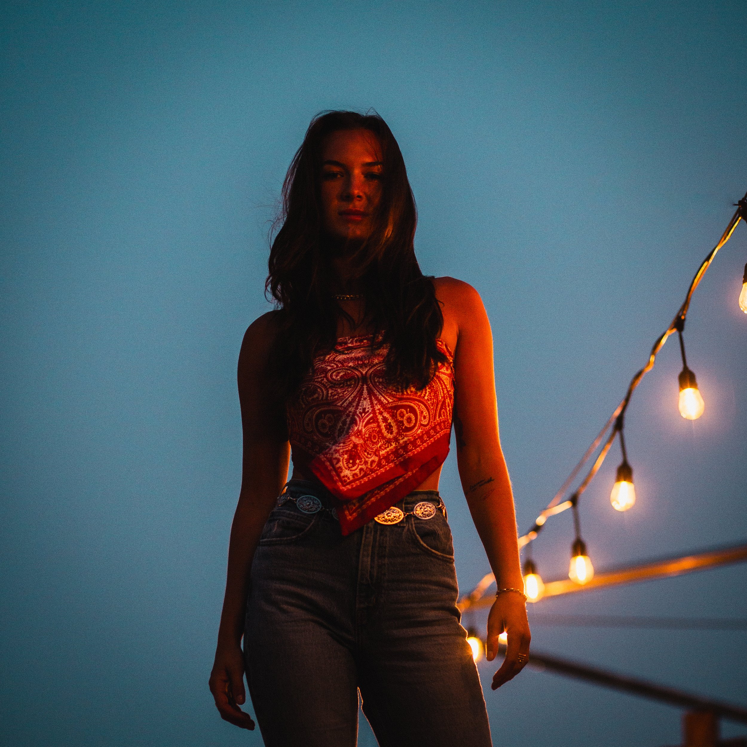 A young woman with long dark hair stands outdoors under a string of glowing lights during dusk, wearing a red bandana-patterned sleeveless top and high-waisted jeans.