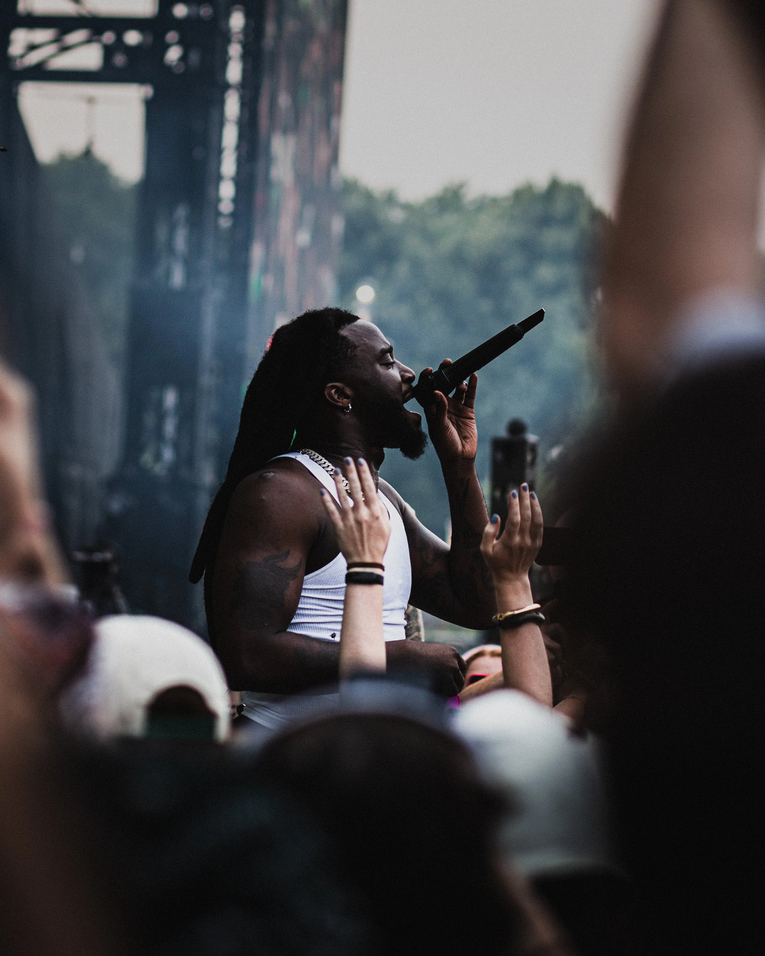 A male performer with long dreadlocks and tattoos singing into a microphone at an outdoor concert, with a crowd in the foreground.