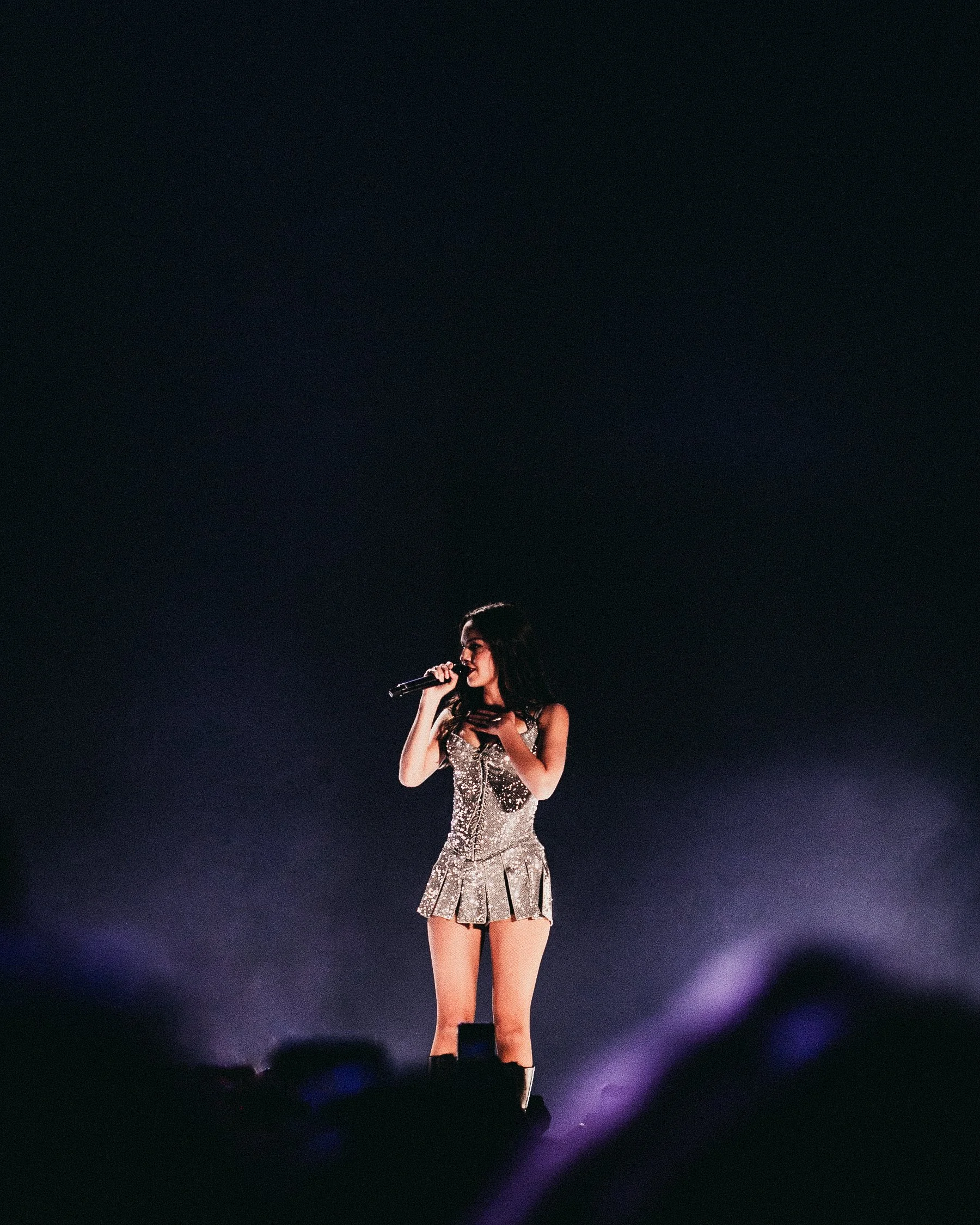 A woman is performing on stage, holding a microphone, wearing a sparkly silver dress with a pleated skirt, and standing in front of a dark background with stage lights.