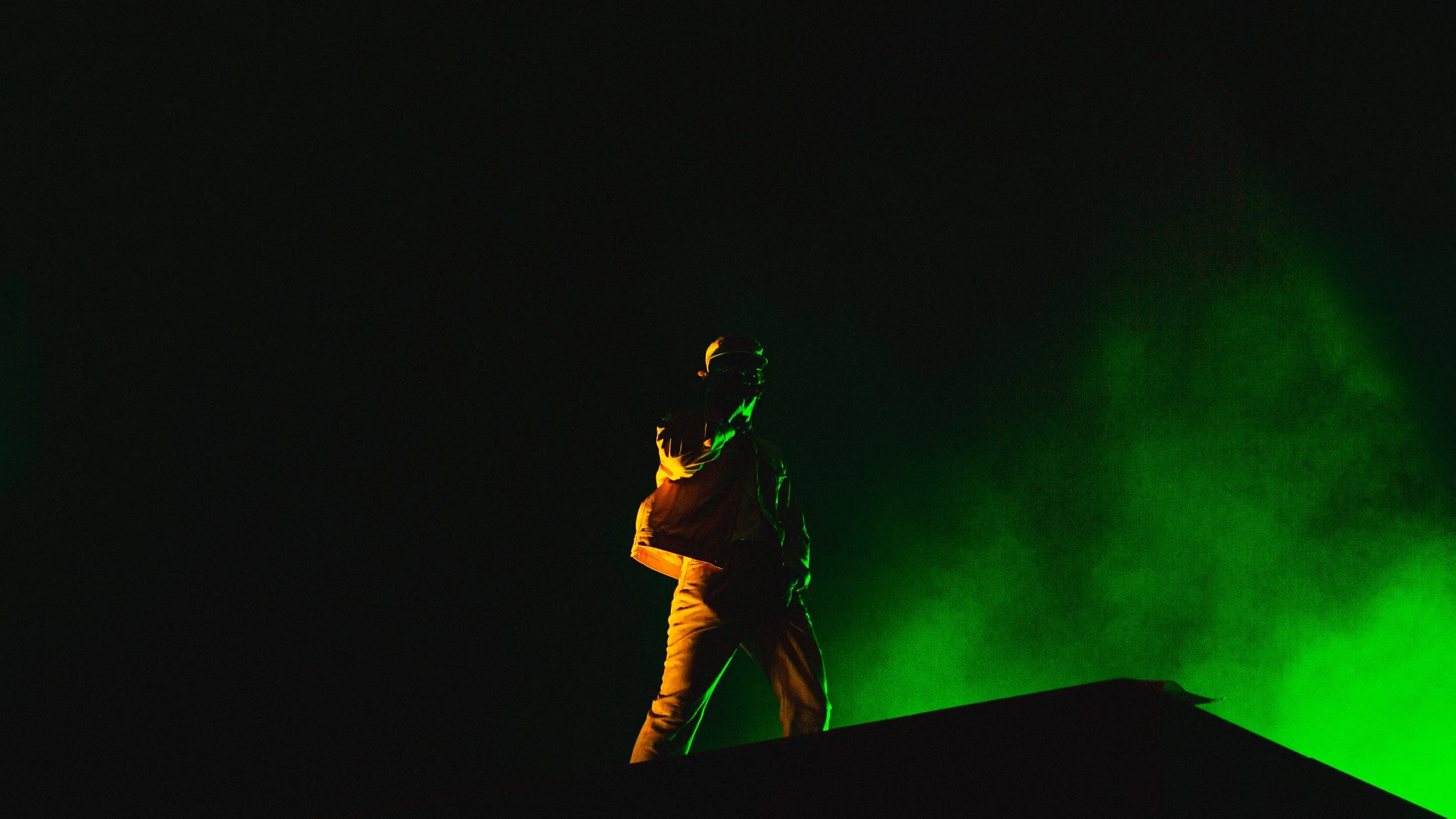 Silhouette of a person standing on a rooftop at night, illuminated by green and yellow lights, with a dark sky in the background.